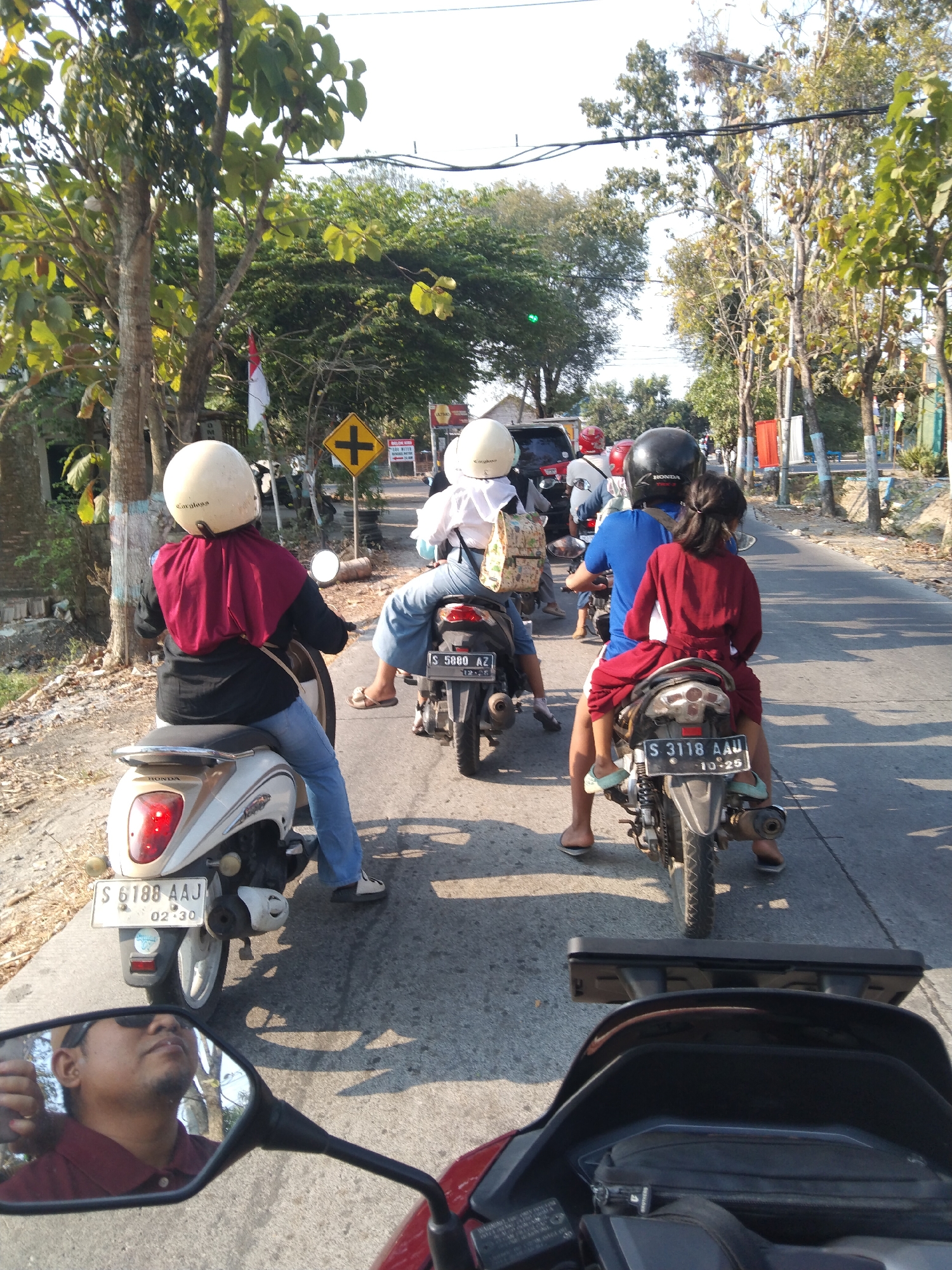 A group of people on motorcycles are traveling on a paved road in Ngraseh, Indonesia. In the foreground, a person is reflected in a motorcycle's mirror, wearing a maroon shirt and sunglasses. Behind them, a white motorcycle is visible with a red taillight. Further down the road, a person in a red headscarf and black shirt is riding a white motorcycle. Next, a person in a white helmet and blue jeans is riding a motorcycle. Behind them, a person in a blue shirt is riding a motorcycle with a young girl in a red dress as a passenger. Another motorcycle is behind them, its rider wearing a black helmet. The road is lined with trees and utility poles. A yellow road sign with a black plus symbol is visible on the right side of the road. The sky is clear and bright.