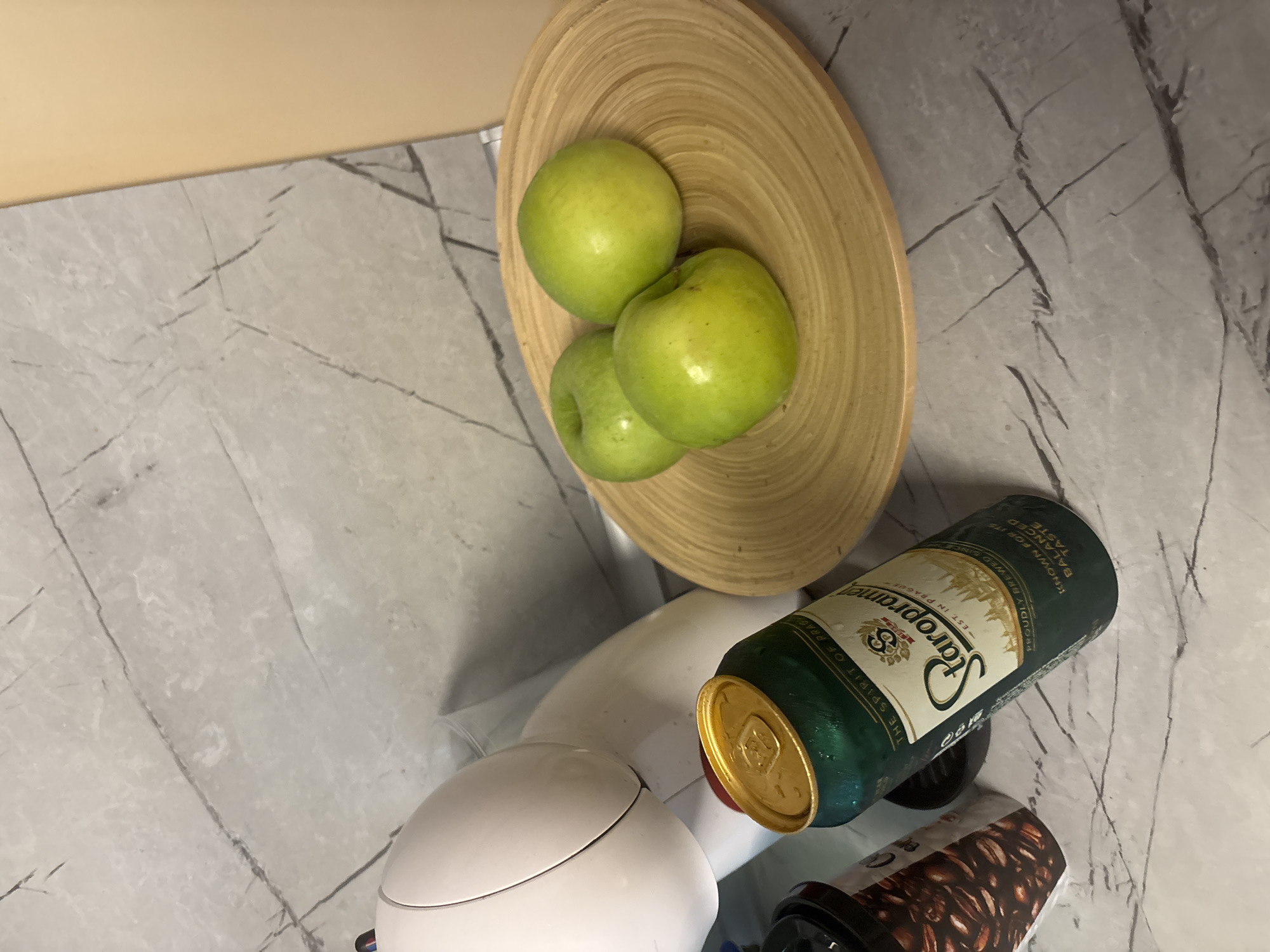 This media file captures a still life composition on a light grey, marbled countertop, suggestive of a kitchen or bar setting. A beige or light yellow wall is visible along the upper left edge of the frame, creating a clean diagonal division with the countertop surface.
The prominent objects include a light brown, circular wooden bowl with distinct concentric grooves, placed centrally towards the top of the image. Inside the bowl are three vibrant green apples.
Below and to the right of the bowl, a dark green beer can with a golden top is prominently displayed. The can appears to be cold, indicated by condensation droplets on its surface. Text visible on the can, also extracted via OCR, includes: "THE SPIRIT OF PRAG," "Staropramen," "EST IN PRAGUE," "PROUDLY BREWED SINCE," "KNOWN FOR Iイン TASTE," and "BALANCED." The can also indicates "5.0% ALC."
Further down, a white, smooth-surfaced appliance, possibly the base of a blender or a coffee maker, is partially visible. To its right, a dark-colored tumbler or bottle featuring a label with a pattern of coffee beans can be seen.
No individuals or discernible activities are taking place. The scene is static, set indoors, with no clear indicators of the time of day or external weather. The location context provided is Varna, Bulgaria.