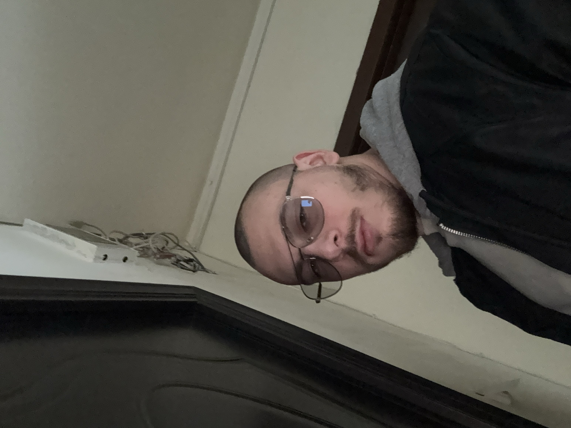 An indoor, headshot-style image, rotated 90 degrees clockwise, features a young man with a shaved head, dark-rimmed glasses, and a short beard. He is wearing a grey hooded sweatshirt under a black zip-up jacket. His expression is neutral as he looks directly towards the camera. Reflections are visible on his glasses.
The background consists primarily of a light beige or off-white wall. In the upper left portion of the image (as originally viewed), a white rectangular object with two small holes is visible, connected to a tangle of white and colored electrical wires and cables. This setup appears to be resting on a ledge or the top of a dark-colored door frame, which occupies the lower part of the image. The overall setting suggests an interior space, possibly a room or hallway, in Stara Zagora, Bulgaria, with some exposed utilities. The lighting is artificial, indicating an indoor environment where the time of day is not discernible. No visible text is present in the image.