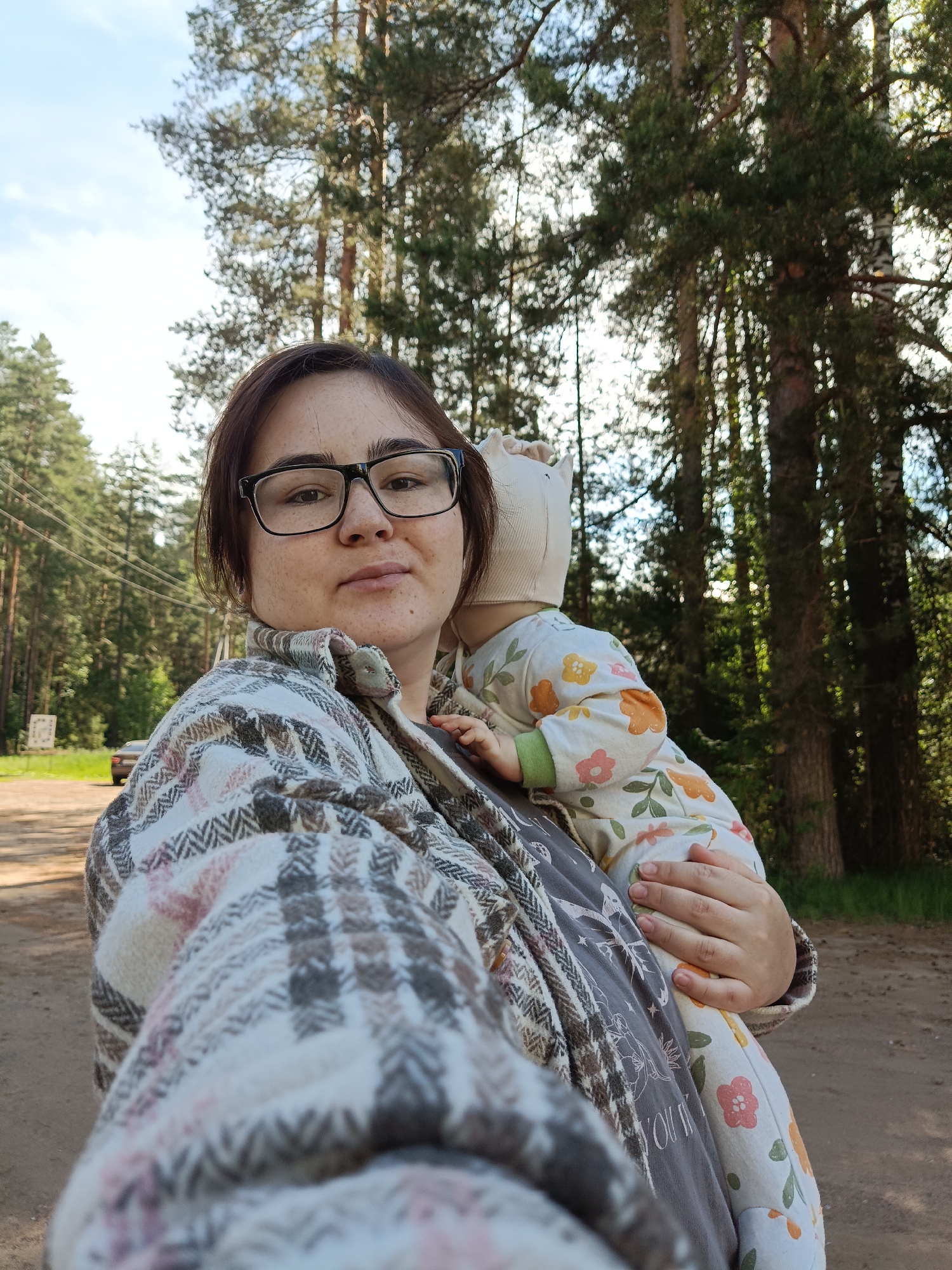 A woman with dark hair and glasses takes a selfie outdoors, holding a baby. She has a neutral expression and visible freckles, wearing a plaid jacket with brown, white, and pinkish hues over a dark t-shirt. The baby, held against her chest, is turned away from the camera, showing only the back of its head and body. The baby wears a light-colored onesie with orange and pink floral patterns and green leaves, and a light bonnet with a small crown-like detail.
They are standing on a dirt or gravel road, flanked on both sides by a dense forest of tall coniferous trees. The sky above is bright and partly cloudy, indicating daytime, likely late morning or afternoon. In the background on the left, a dark car is partially visible on the road, along with utility poles and wires, and an unreadable road sign. The setting suggests a natural, rural or semi-rural location, consistent with the Noviy, Russia context. The scene captures a casual outdoor moment, possibly during a walk in the woods. No clear text is legible on any objects in the image.