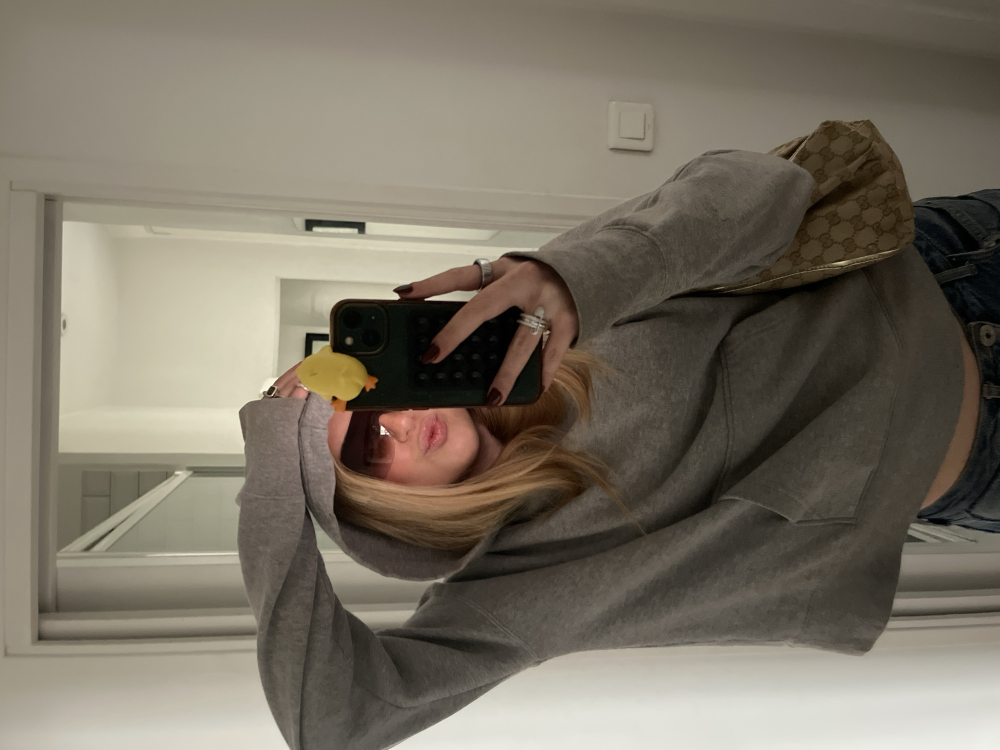 A person wearing a gray hoodie and jeans takes a selfie in a bathroom mirror. The person is holding a smartphone with a yellow rubber ducky attached to it. A tan handbag is slung over their shoulder. The bathroom has white walls and a white door frame. There is a light switch on the wall. The location is not discernible as being Paris, France, based solely on the visual evidence.