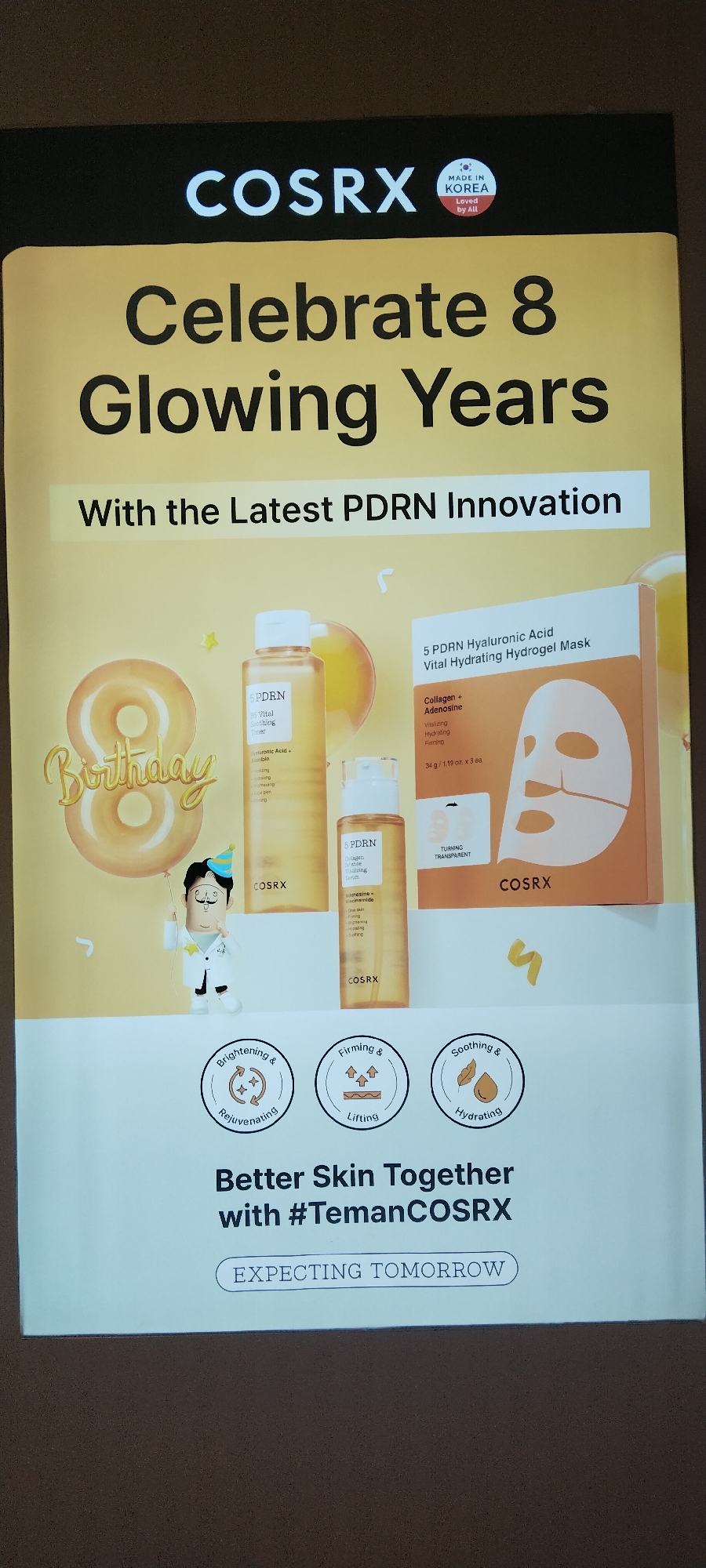 The image is a promotional poster for COSRX, a Korean skincare brand, celebrating its 8th anniversary. The poster features the COSRX logo prominently at the top, along with a "Made in Korea" emblem. The central message, "Celebrate 8 Glowing Years," is displayed in large font, followed by the tagline, "With the Latest PDRN Innovation."
Visually, the poster is dominated by a bright yellow background. To the left, a large, golden balloon shaped like the number 8 is visible, with the word "Birthday" written in a stylized font below it. A small cartoon character, appearing to be a doctor or scientist, stands to the left of the product display, holding a balloon string.
In the center and right of the poster, COSRX skincare products are showcased. There is a bottle of toner and a smaller bottle of serum, both labeled "5 PDRN." A box of "5 PDRN Hyaluronic Acid Vital Hydrating Hydrogel Mask" is also prominently displayed. The products appear to be in amber-colored glass bottles and packaging.
Below the product display, three circular icons illustrate the benefits of the products: "Brightening & Rejuvenating," "Firming & Lifting," and "Soothing & Hydrating."
The bottom of the poster features the slogan, "Better Skin Together with #TemanCOSRX," and a call to action, "EXPECTING TOMORROW." The overall design is clean, modern, and aims to convey a sense of luxury and innovation in skincare.
The image's context as a promotional poster suggests it might be displayed in a retail store, a shopping mall, or an event venue. Without further information or contextual clues within the image itself, it is impossible to pinpoint its specific location within the city of Semarang, Indonesia.
