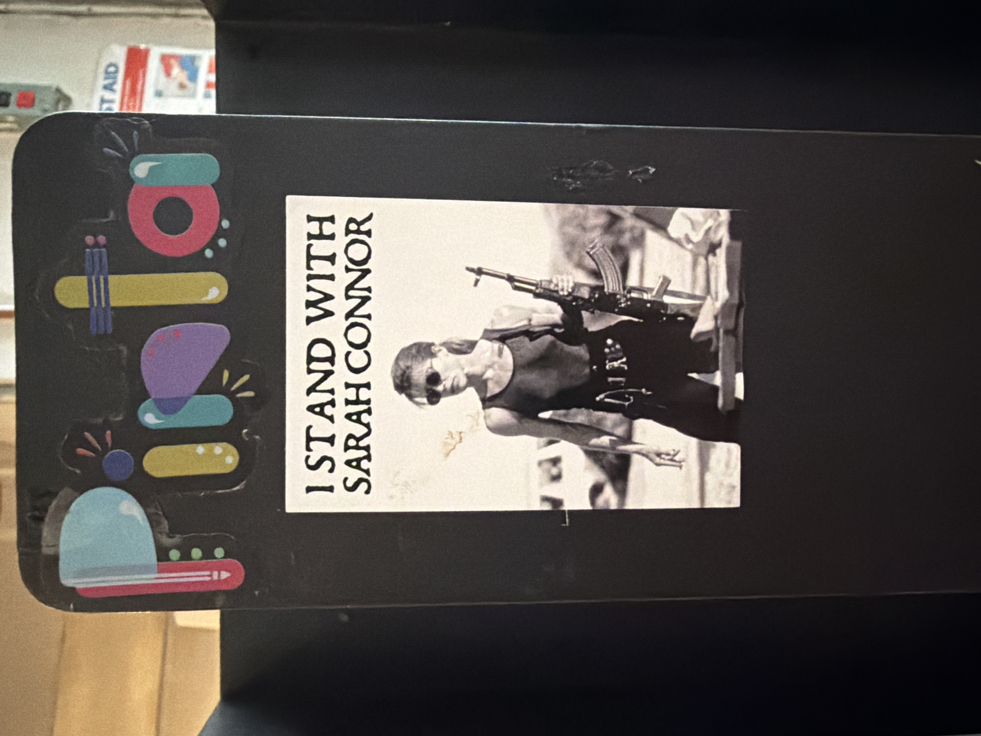 The image shows a rectangular sticker with the text "I STAND WITH SARAH CONNOR" beside a black and white image of a woman holding a rifle. The sticker is affixed to a dark, possibly metal, surface. To the left of the sticker is a design composed of stacked colorful shapes: turquoise, pink, yellow, purple, and light blue with red accents. A first aid kit is partially visible in the background. The location appears to be indoors. There are no specific landmarks to identify it as being in the city of New York.