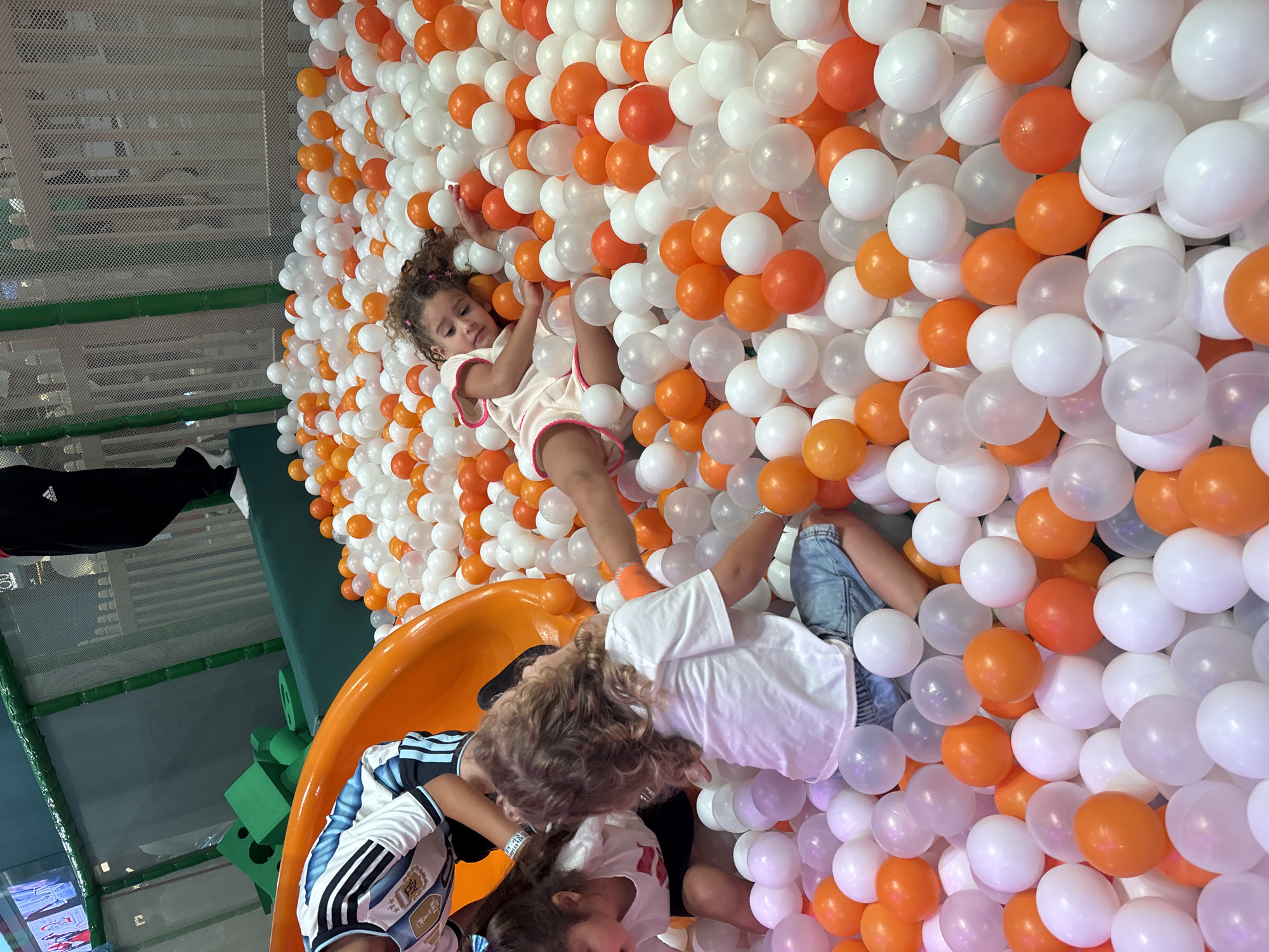 A group of young children are playing in a ball pit filled with numerous white and orange plastic balls. One child, wearing a white and blue striped soccer jersey, is partially visible in the upper left of the frame, near an orange slide. Another child, a girl with curly brown hair, lies on her side in the middle of the ball pit, looking towards the camera. A third child, with blond curly hair and wearing a white t-shirt and denim shorts, is seated in the ball pit, facing away from the camera. In the background, a person in black pants and white shoes stands near a green and white netted barrier. The environment appears to be an indoor playground in Dubai, United Arab Emirates.