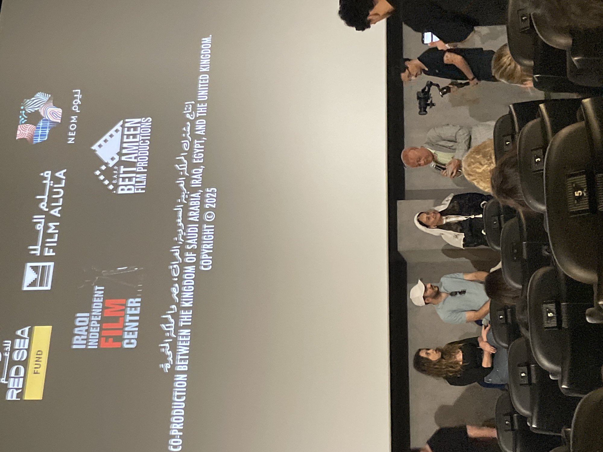 The image depicts a darkened movie theater with several rows of seats occupied by an audience. The screen displays the production credits for a film, indicating it is a co-production between the Kingdom of Saudi Arabia, Iraq, Egypt, and the United Kingdom. The credits also mention organizations like the "Red Sea Fund," "Iraqi Independent Film Center," "Film AlUla," and "Beit Ameen Film Productions," with a copyright date of 2025. Some people in the audience appear to be filming or taking pictures of the screen, suggesting a premiere or special screening event. The image offers no information on a city in Barcelona, Spain.
