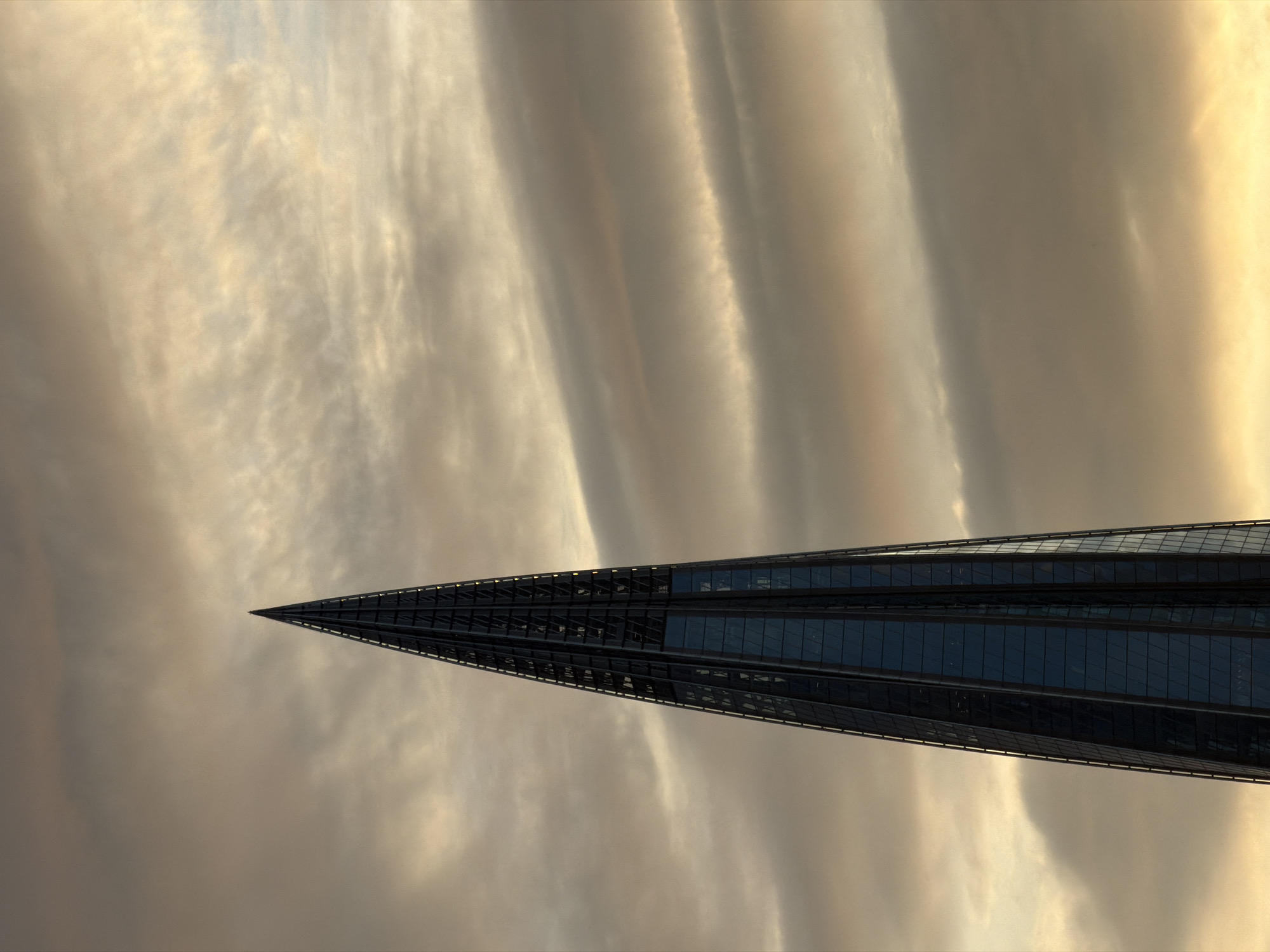 The image presents a modern, glass-clad skyscraper, likely the Lakhta Center in Saint Petersburg, Russia, sharply ascending into a dramatic sky. The building occupies the right side of the frame, tapering elegantly to a very sharp, needle-like spire that reaches towards the top of the image. Its facade is composed of dark, reflective glass panels, giving it a sleek and sophisticated appearance.
The sky, which fills the majority of the frame, is a canvas of soft, undulating clouds. These clouds display a palette of warm golden, soft yellow, and various shades of muted gray, suggesting either the soft light of sunrise or sunset. Lighter, brighter streaks of clouds are interspersed with darker, more shadowed areas, creating a textured and dynamic atmospheric backdrop.
There are no people or discernible activities taking place, placing the sole focus on the architectural marvel against the natural sky. The lighting is diffused and atmospheric, highlighting the contours of the clouds and the reflective surfaces of the building. The image conveys a sense of grandeur, modernity, and the impressive scale of human construction set against a vast sky. No text is visible in the image.
