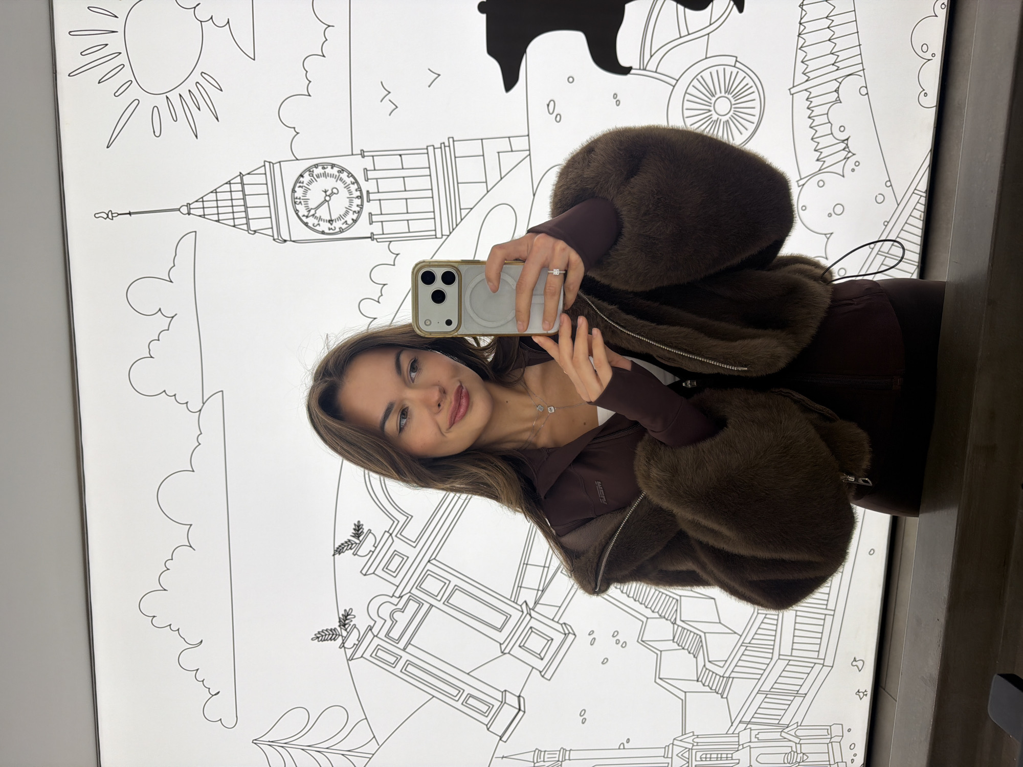 A selfie shows a young woman with brown hair and fair skin holding a cell phone. She is wearing a brown jacket with fur lining. She is standing in front of a wall depicting a drawing of London, which includes drawings of Big Ben, the Houses of Parliament, and a silhouette of a bear riding a bicycle. The location is not in Krasnoyarsk, Russia.