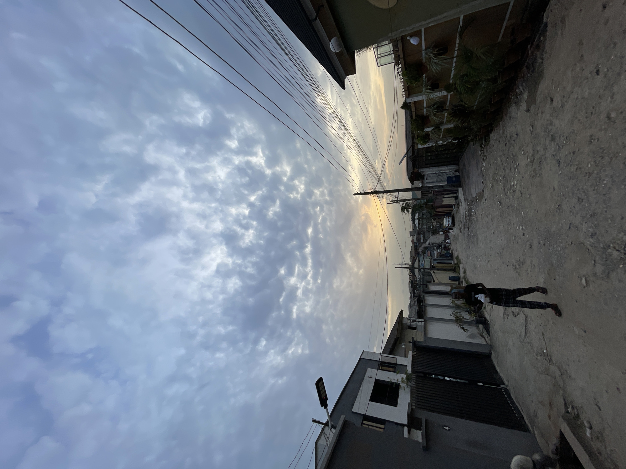 The image shows a street in Isheri, Nigeria. The view is tilted to the side, capturing a cloudy sky overhead with power lines stretching across. Buildings line both sides of the street, with a two-story building on the right featuring plants on its balcony and a modern, gray building with a black metal gate on the left. A person is walking down the street on the right side. The road surface appears to be dirt and gravel.