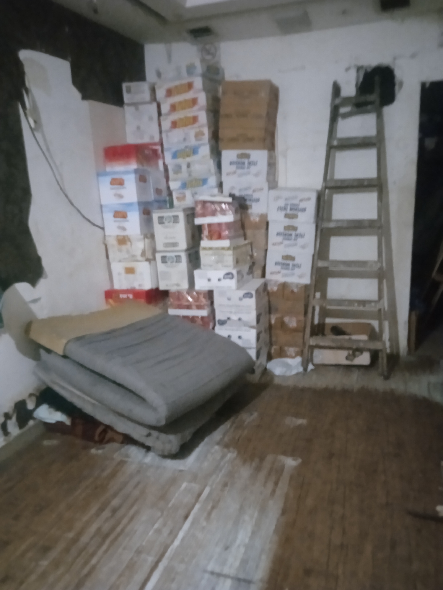 The image depicts an indoor scene in a room, likely a storage area or a shop, located in the city of Gaza, Palestinian Territories. A tall wooden ladder leans against the right wall, with several stacked cardboard boxes of various sizes and colors filling the space. Some boxes are white with red and blue text, while others are brown. In the foreground, a gray folded mattress or sleeping mat rests on a flat surface, with some darker fabric underneath. The floor is made of wooden planks and appears somewhat worn. The walls are white, with some visible damage and discoloration. The lighting is dim, suggesting either low ambient light or a poor quality camera. There are no people visible in the image. The scene conveys a sense of disarray and storage.