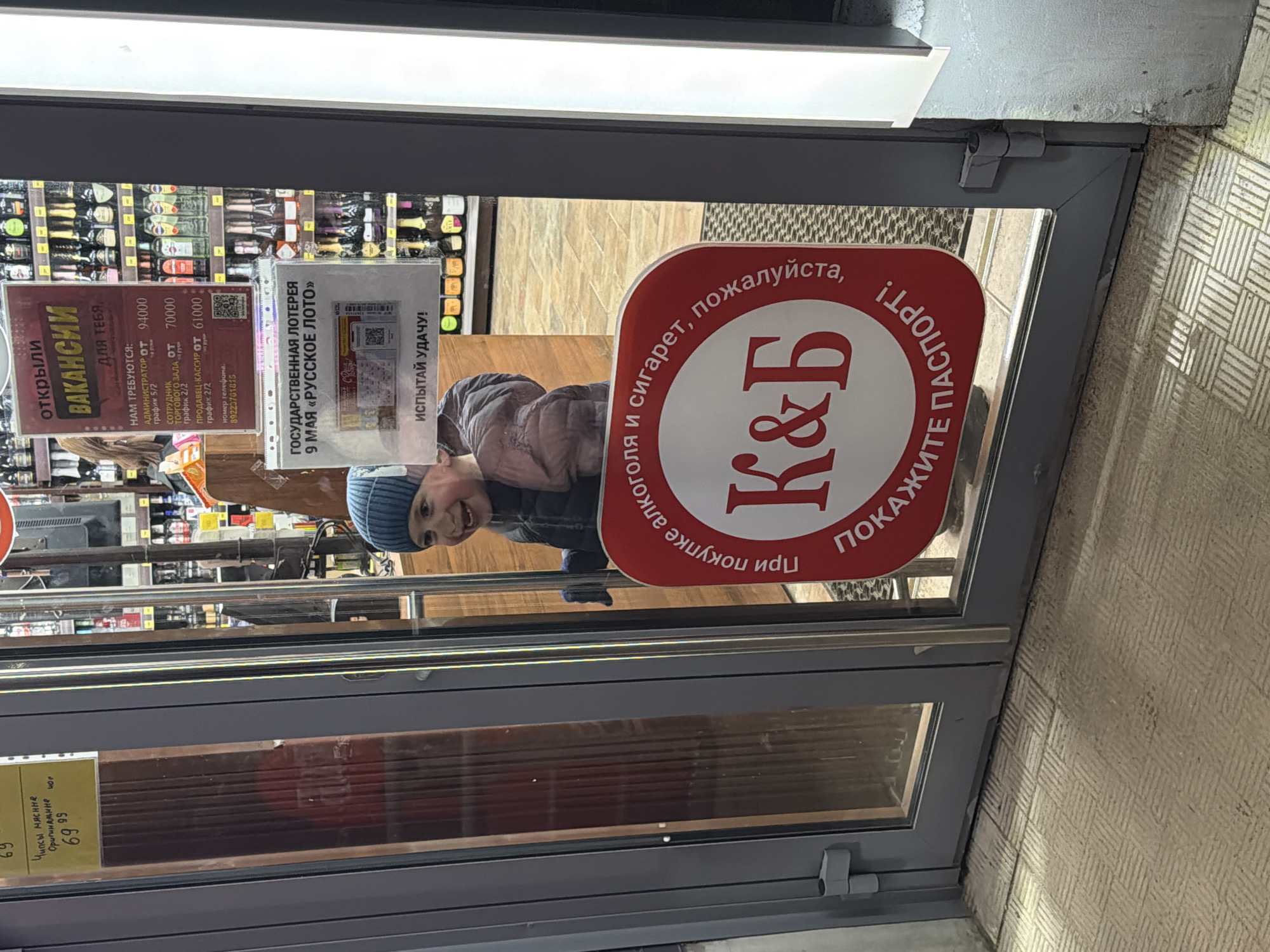 A young child, wearing a blue beanie and a jacket, looks through the glass door of a store, smiling broadly. The image is taken from outside, capturing a view into a retail establishment in Chaikovsky, Russia. The store's interior shows shelves stocked with numerous bottles, likely alcohol, indicating it might be a liquor store or a supermarket section.
Attached to the glass door are several signs. A prominent red circular sign features the "K&B" logo and text in Russian: "При покупке алкоголя и сигарет, пожалуйста, ПОКАЖИТЕ ПАСПОРТ!" (When purchasing alcohol and cigarettes, please SHOW PASSPORT!). To the left, a "ВАКАНСИИ" (VACANCIES) sign advertises job openings for an Administrator (from 94000), Sales Floor Employee (from 70000), and Salesperson-Cashier (from 61000), along with a phone number. To the right, a sign promotes "ГОСУДАРСТВЕННАЯ ЛОТЕРЕЯ 9 МАЯ «РУССКОЕ ЛОТО»" (STATE LOTTERY MAY 9 "RUSSKOE LOTO") with the phrase "ИСПЫТАЙ УДАЧУ!" (TRY YOUR LUCK!). A small price tag for "Чипсы мясные Оригинальные" (Meat chips Original) at "69 99" is visible on the bottom left of the door. The child's cheerful expression is a notable detail, contrasting with the commercial information displayed on the door. The scene appears to be during daytime.
