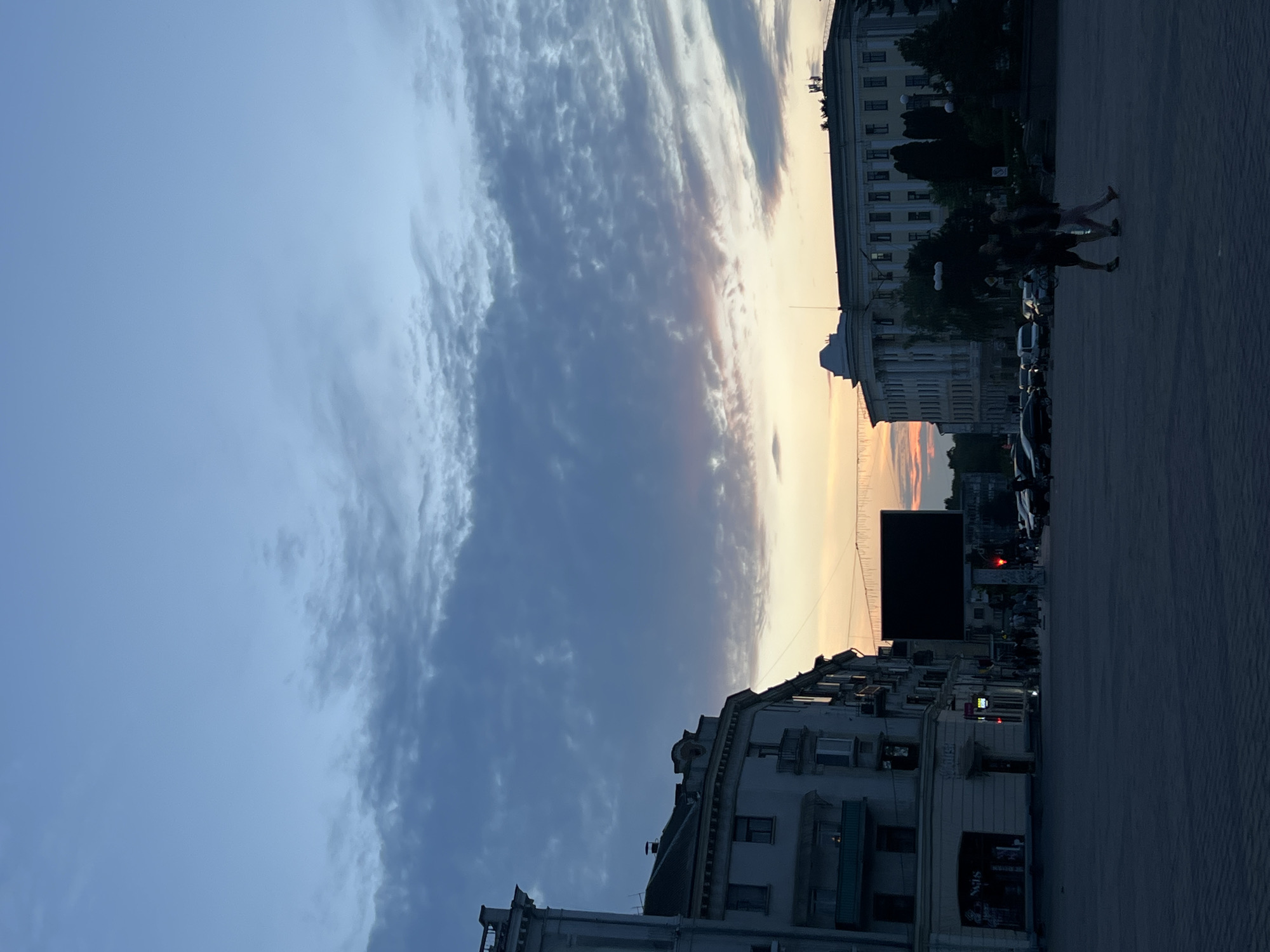 The image captures an urban street scene at dusk or twilight, oriented vertically but depicting a horizontal view of a city street. The sky dominates the upper portion, transitioning from a soft blue on the left to warm oranges and yellows on the right, indicative of a sunset. Streaky, textured clouds are prominent against the fading light.
Along the street, buildings of various sizes and architectural styles line both sides. On the right, a large, lighter-colored building with numerous windows and decorative elements is visible, partially silhouetted. On the left, another multi-story building appears darker, with visible windows and what might be balconies. Further down the street, more structures are obscured by shadows.
Several people are walking on the right side of the street in the midground, their figures mostly silhouetted against the ambient light. Cars are parked or moving along the street, also in shadow. A large, dark rectangular billboard or screen stands out in the distance on the left. Faint red lights, possibly traffic signals or building lights, are discernible. Some string lights or wires are strung across the street near the horizon line.
The overall lighting suggests the transition from day to night, with natural light from the setting sun casting long shadows and creating silhouettes, while artificial lights from buildings and street furniture begin to emerge. Text is barely visible on a sign on the lower left building, possibly reading "SIGN" and another word. Another faint sign further down looks like "BAR".