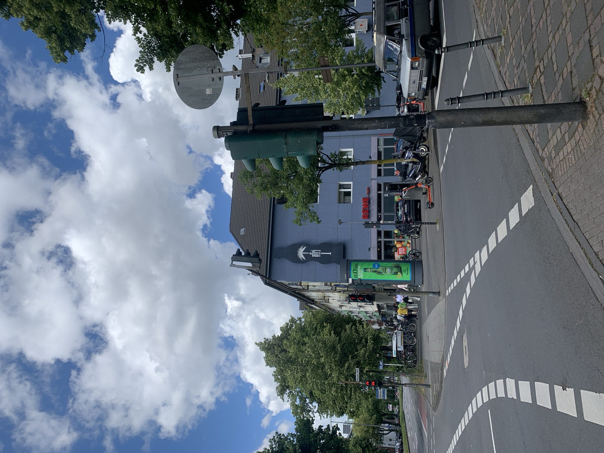 The image captures a vibrant street scene on a clear, partly cloudy day. The sky is bright blue with scattered white, fluffy clouds. Lush green trees line parts of the street and frame the top of the image.
In the foreground, an asphalt road with distinct white dashed lane markings curves, suggesting an intersection or turn. A paved sidewalk runs alongside the road, where several dark bollards are visible. Numerous vehicles are present, including a white van with the license plate "WFH B241", parked cars, and a cluster of electric scooters (including orange and lime green models) and bicycles.
Several buildings line the street. A prominent blue-grey building features a large, stylized black bottle-shaped sign with white text, possibly reading "ARI CANAL". Below this, a section of a building displays a red "REWE" logo, which also appears on a green advertising board further down the street, promoting a "0,0 Alc." product. Multiple traffic lights are visible; some show a green signal, while others display red, indicating active traffic management. A street sign attached to a pole reads "Neuer Wall". A few people are seen on the sidewalk, appearing to be walking or waiting near the buildings. The overall scene depicts a busy urban environment during daylight hours.