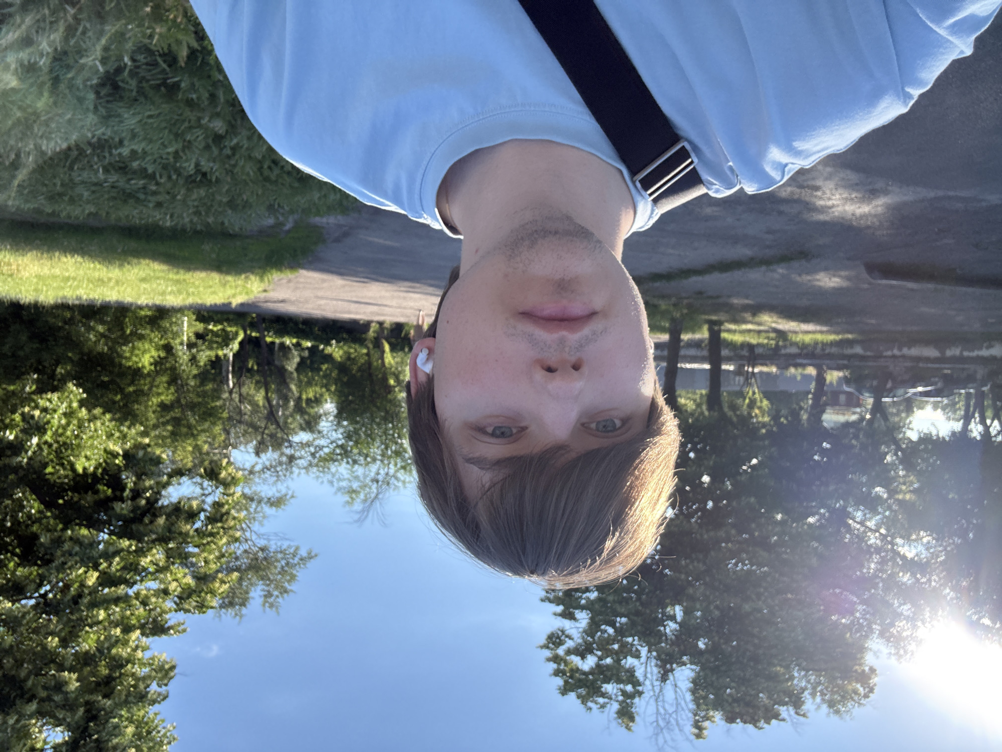 The image is a selfie of a young white male with brown hair, fair skin, and blue eyes, taken outdoors on a sunny day. He is wearing a light blue t-shirt and a black strap across his shoulder, with white wireless earbuds in his ears. He appears to be in a park or residential area, with trees and green grass visible in the background. A paved path and a few parked cars can also be seen. The overall impression is a casual, everyday scene. The location is specified as Dnipro, Ukraine.