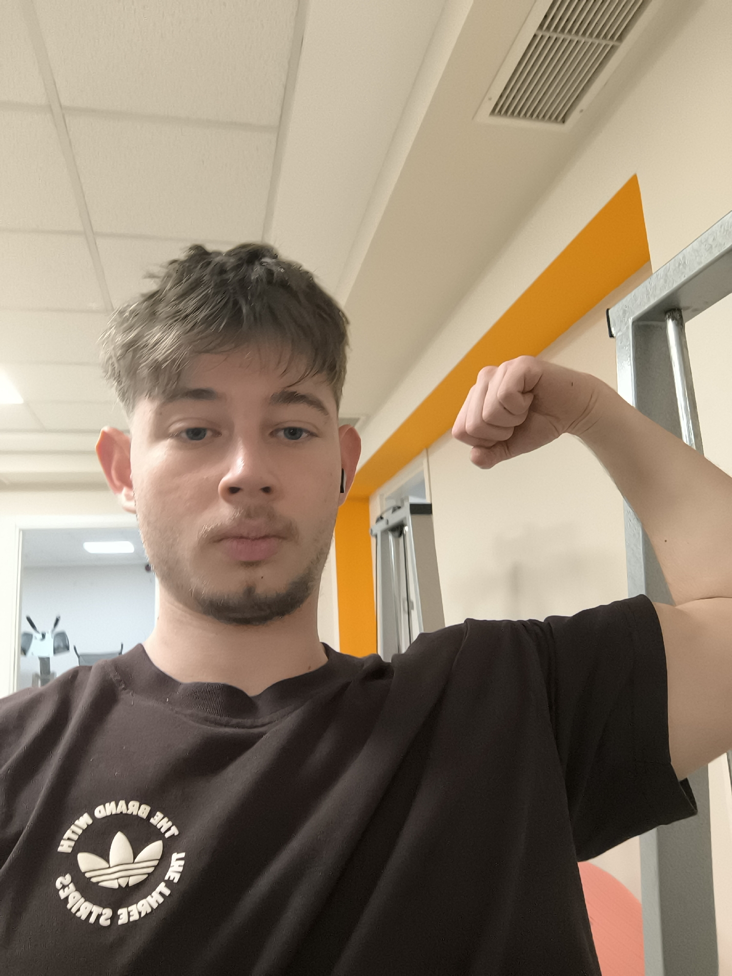 A young man poses for a selfie in what appears to be a gym or fitness center. He is wearing a black Adidas t-shirt and flexing his right bicep. The background includes gym equipment, orange wall accents, and a metal structure. The lighting is bright and artificial, common in indoor fitness facilities. The location details (city of Sofia, Bulgaria) are not visually discernible from the image itself.