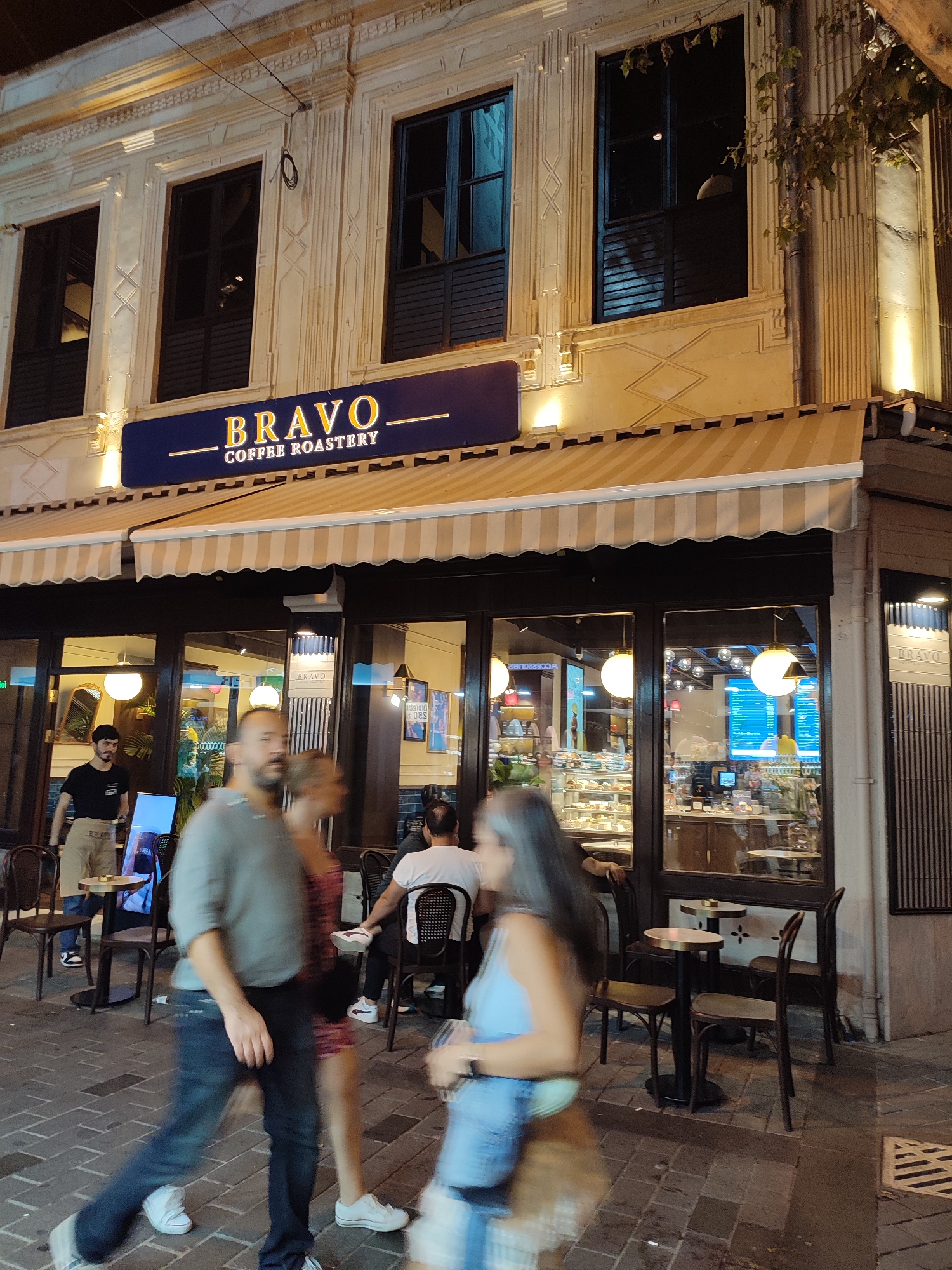 The image captures the exterior of a Bravo Coffee Roastery located in Kadıköy, Turkey, at night. The building facade features a dark blue awning with the coffee shop's name and "Coffee Roastery" in white lettering. Above the awning, there are three large windows with dark shutters, set within decorative architectural elements. The street in front of the cafe is paved with brickwork. Several people are visible, some blurred due to motion, walking on the sidewalk or seated at tables outside. One person, a man in a gray shirt and dark jeans, is walking towards the viewer. Another person, a woman in a light blue top, is also walking. Inside the cafe, through large glass windows, displays of pastries and coffee-making equipment are visible. Warm lighting illuminates the interior and the cafe's exterior.