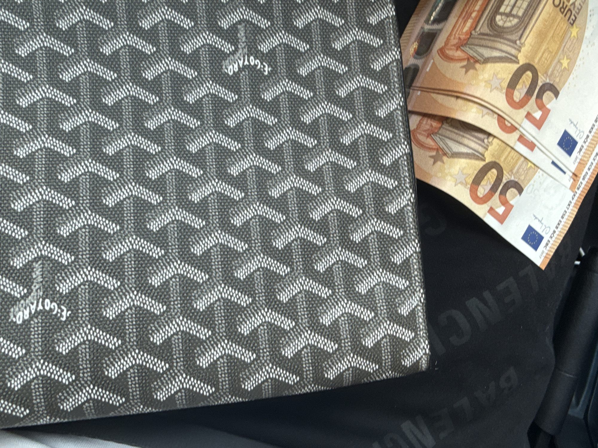 The scene depicts a close-up, slightly overhead view of luxury accessories, captured in the city of Plovdiv, Bulgaria. Dominating the upper and central portion of the frame is a dark grey item, likely a bag or wallet, featuring the distinctive interlocking chevron-like pattern composed of white dots, characteristic of the Goyard brand. The text "E. GOYARD" is discernible in white within this pattern towards the top right. Below this item, a black fabric surface is visible, bearing embossed or printed text. Partially legible letters include "BALEN" on the left and "LENC" on the right, strongly suggesting the brand name "BALENCIAGA". Nestled in the bottom right corner, partially obscured by the Goyard item, are two 50 Euro banknotes. They are folded and overlapping, clearly displaying the number "50" in orange and the blue and yellow European Union flag emblem. No individuals, actions, or direct interactions are visible. The environment appears to be an interior setting, possibly a car or a personal space, given the close proximity and arrangement of the items.