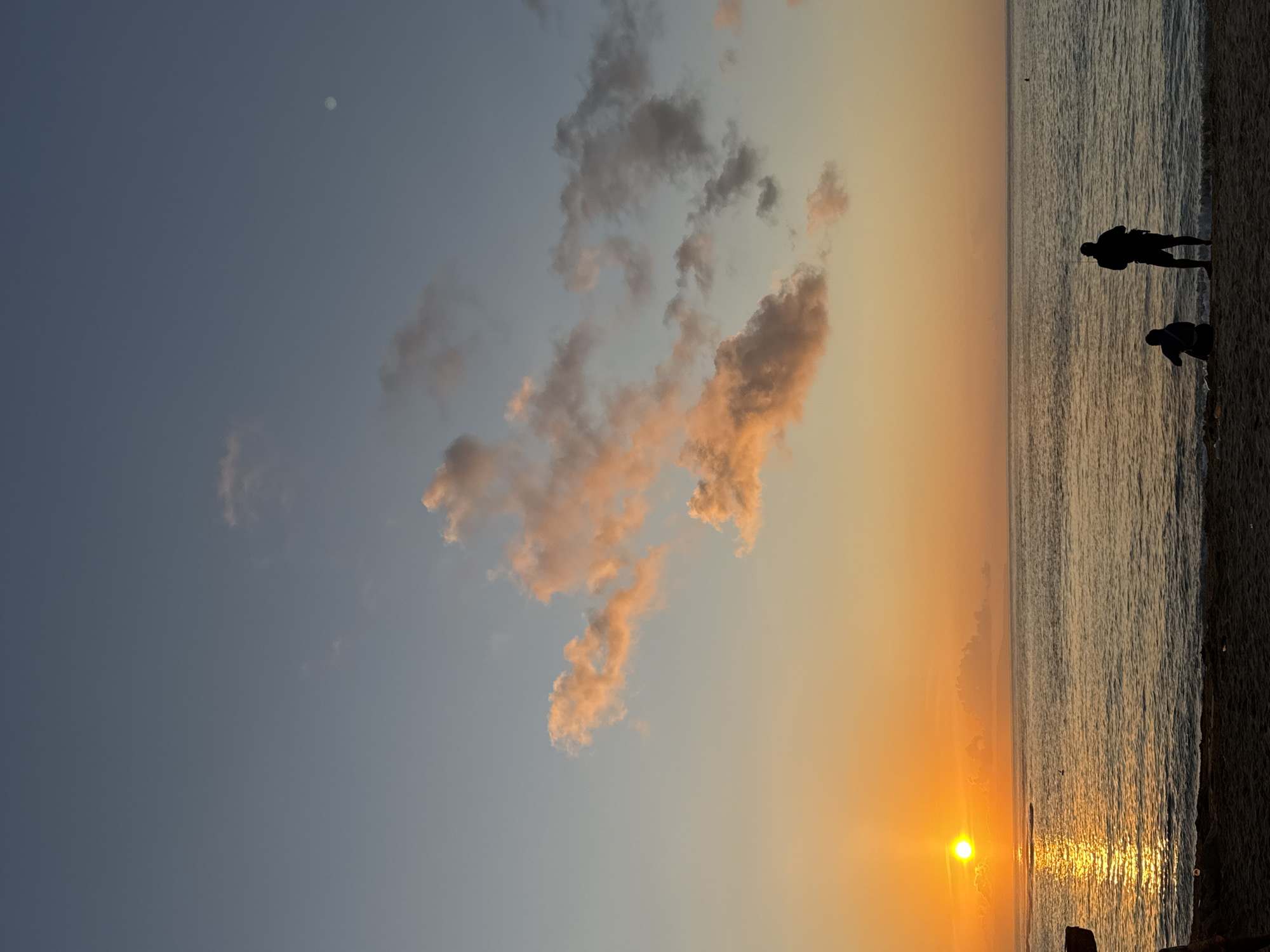 The image shows a beach scene, likely at sunset. The location is Kuta Selatan, Indonesia. The sky transitions from blue to orange, with the sun low on the horizon, casting a bright reflection on the water. There are scattered clouds, tinged with orange and pink hues. Two silhouetted figures stand at the shoreline. The sea is calm with small waves. The image is vertically oriented.