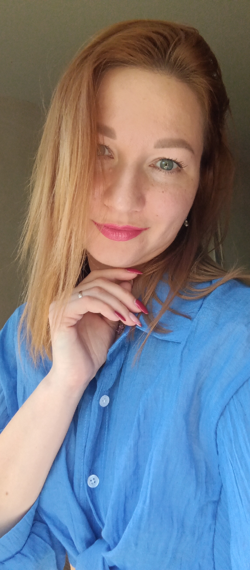 A woman with light reddish-brown hair and green eyes, wearing a blue button-down shirt, poses in front of a gray wall. She is resting her chin on her hand, showing a ring on her finger and bright pink lipstick. Her fingernails are painted red. The location is Unknown, Unknown.
