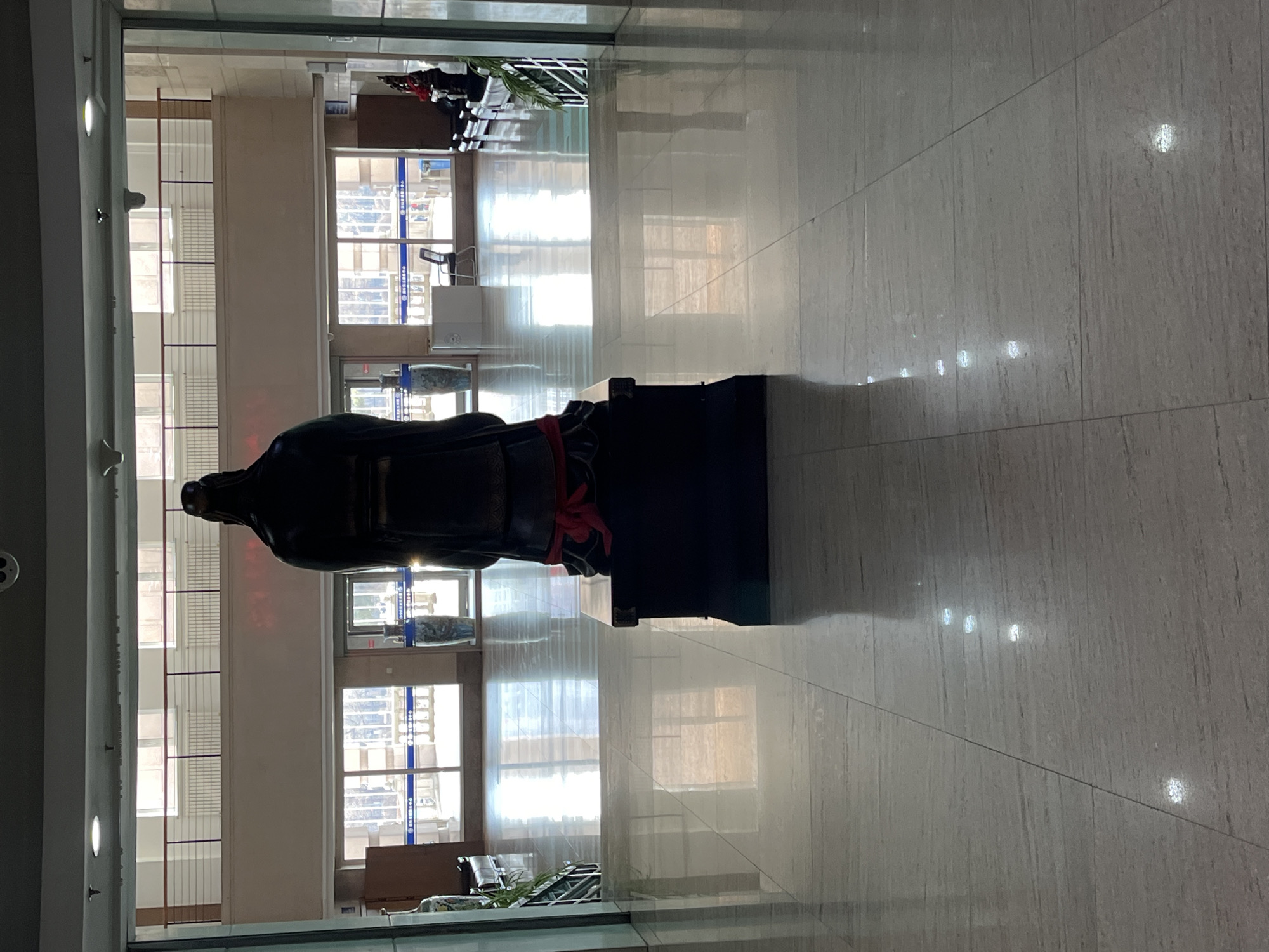 The image depicts the interior of a building, likely a public space in Changchun City, China. A statue stands prominently in the foreground, appearing to be a historical figure in traditional attire, with a red sash or belt around the waist. The statue is placed on a dark-colored base. The background reveals a bright, open area with large windows allowing natural light to illuminate the space. Through the windows, glimpses of the outside world are visible. The floor is tiled with a light-colored, glossy material, reflecting light. The overall impression is that of a modern, well-maintained public building with a cultural artifact as a centerpiece. There are no people visible.