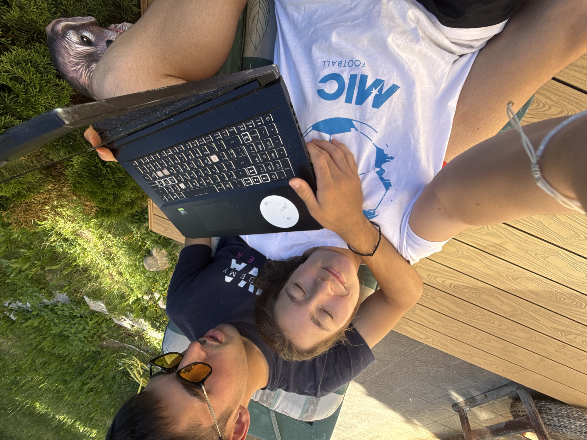 A young couple is lying down outdoors, possibly on a deck or patio, with a laptop in front of them. The woman is leaning her head on the man's shoulder, with her eyes closed and a peaceful expression. The man is wearing sunglasses and looking towards the camera. The scene appears to be set in Sofia, Bulgaria, during a sunny day, suggesting it's likely summer.
The man is wearing a dark blue t-shirt with white lettering that appears to say "ACADEMY". The woman is wearing a white t-shirt with blue graphics. Visible text on the woman's t-shirt includes "MIC" and "FOOTBALL" in blue lettering, along with a graphic of a soccer ball. The overall mood is relaxed and intimate, with the couple enjoying each other's company in an outdoor setting. The background includes greenery, suggesting a garden or park-like environment, and a wooden deck.