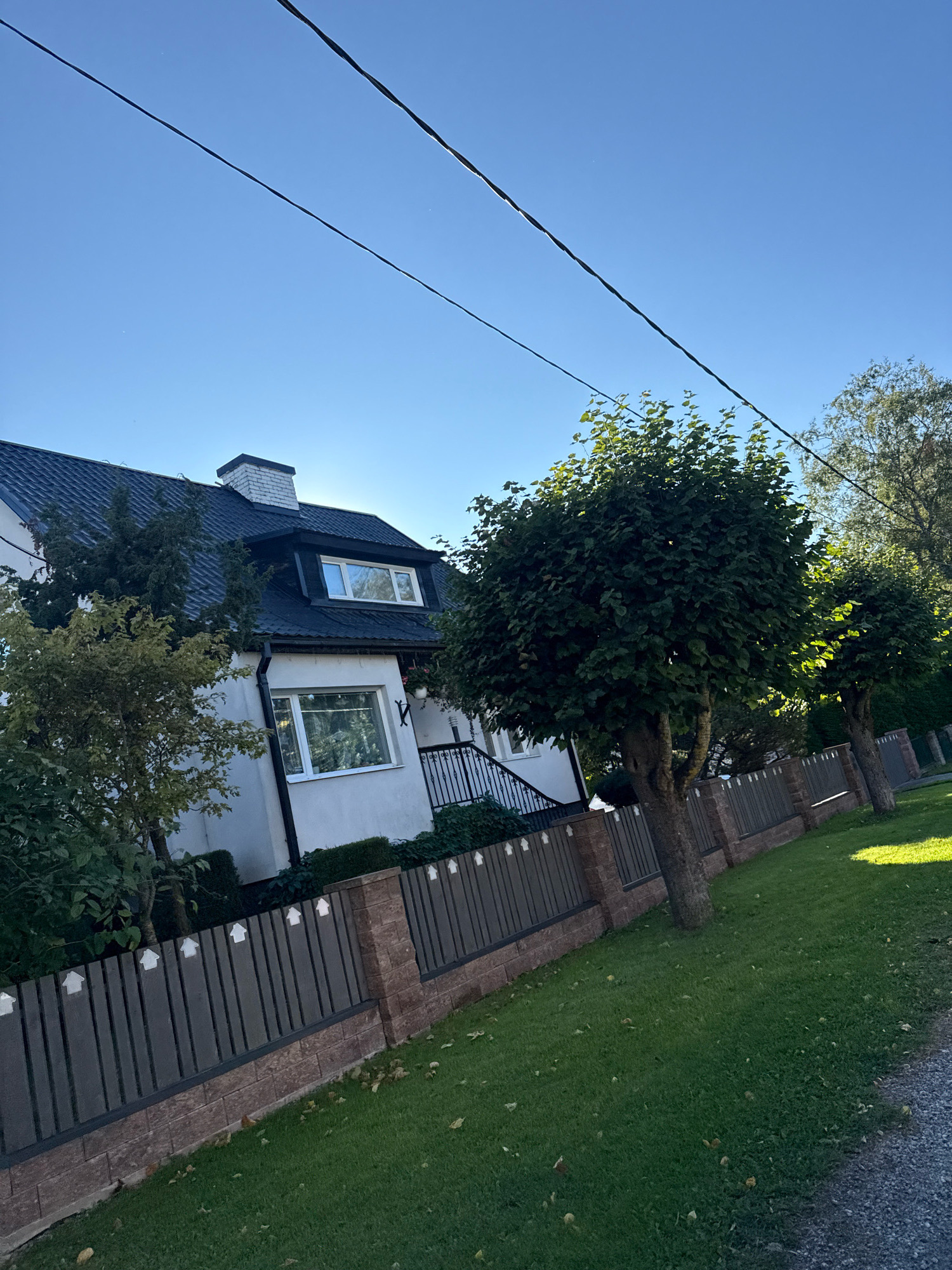 The image depicts a residential property in Tallinn, Estonia, on a clear day. The scene is dominated by a two-story house with a dark roof, white walls, and several windows. A brick chimney with white detailing sits atop the roof. In front of the house, a dark-colored fence with decorative elements separates the property from the street. Two well-maintained trees with full green canopies stand along the fence line, casting shadows on the manicured lawn. The sky is a vibrant blue, with power lines visible stretching across the upper portion of the frame. The overall impression is one of a tidy, well-kept suburban home.