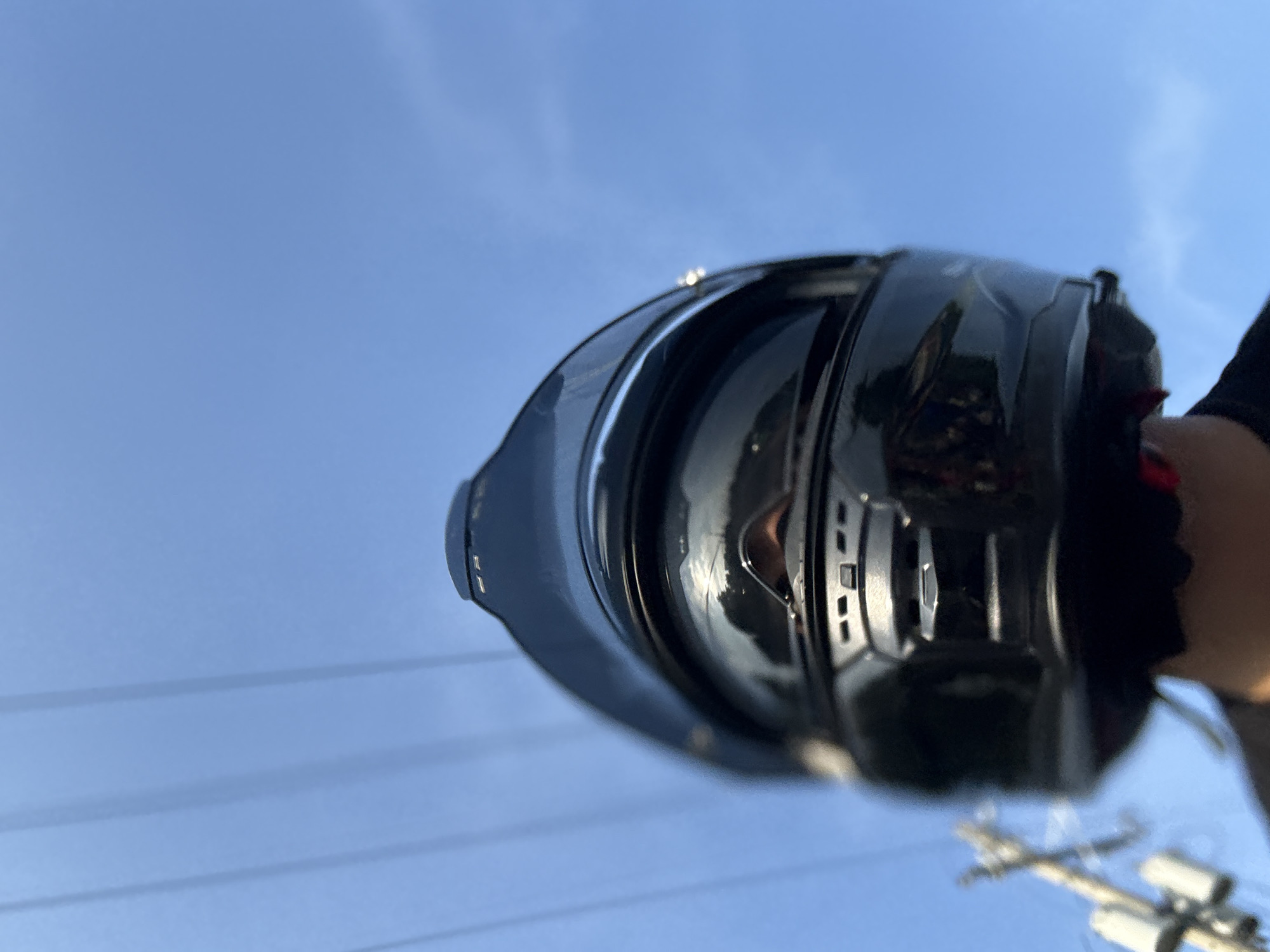 The image shows a close-up, low-angle shot of a person wearing a black motorcycle helmet against a bright blue sky with faint power lines visible in the background. The helmet has a reflective surface that shows a distorted reflection of the person's face and possibly surrounding foliage. The foreground is dominated by the back and top of the helmet, with a small portion of the person's shoulders and neck visible at the bottom. To the left, a blurred power pole with insulators is also present. The scene is likely outdoors, possibly on a road or in an open area within or near Davao City, Philippines, given the presence of power lines and the general outdoor ambiance under a clear sky.
