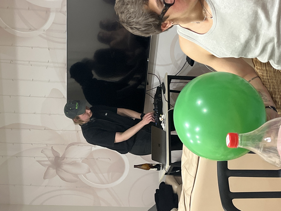 The scene depicts an indoor setting in Заозерное Снт, Russia. In the central portion of the image, a person with light-colored, shoulder-length hair, wearing a dark green baseball cap with "BUTTER" embroidered in light green, a black short-sleeved t-shirt, and headphones around their neck, stands leaning over a black rectangular table. This individual is actively manipulating a black DJ controller with both hands. To the left of the controller, a silver-colored laptop is open on the table, and a brown glass bottle is also present.
Behind the DJ setup, a large, flat-screen television is mounted on a light-colored wall. The television screen is dark and reflective, showing the silhouettes of several indistinct human figures. The wall itself features large, subtle, lighter-toned floral patterns and multiple vertical string-like decorative elements.
To the right, in the foreground, a second individual is partially visible, seated in a wicker chair. This person has short, dark, curly hair, wears dark-rimmed glasses, a grey ribbed tank top, multiple ear piercings, and a silver necklace. A large, bright green spherical balloon occupies the mid-foreground, partially obscuring this individual and a clear plastic bottle with a red cap, which is positioned closer to the viewer. The room is moderately lit.