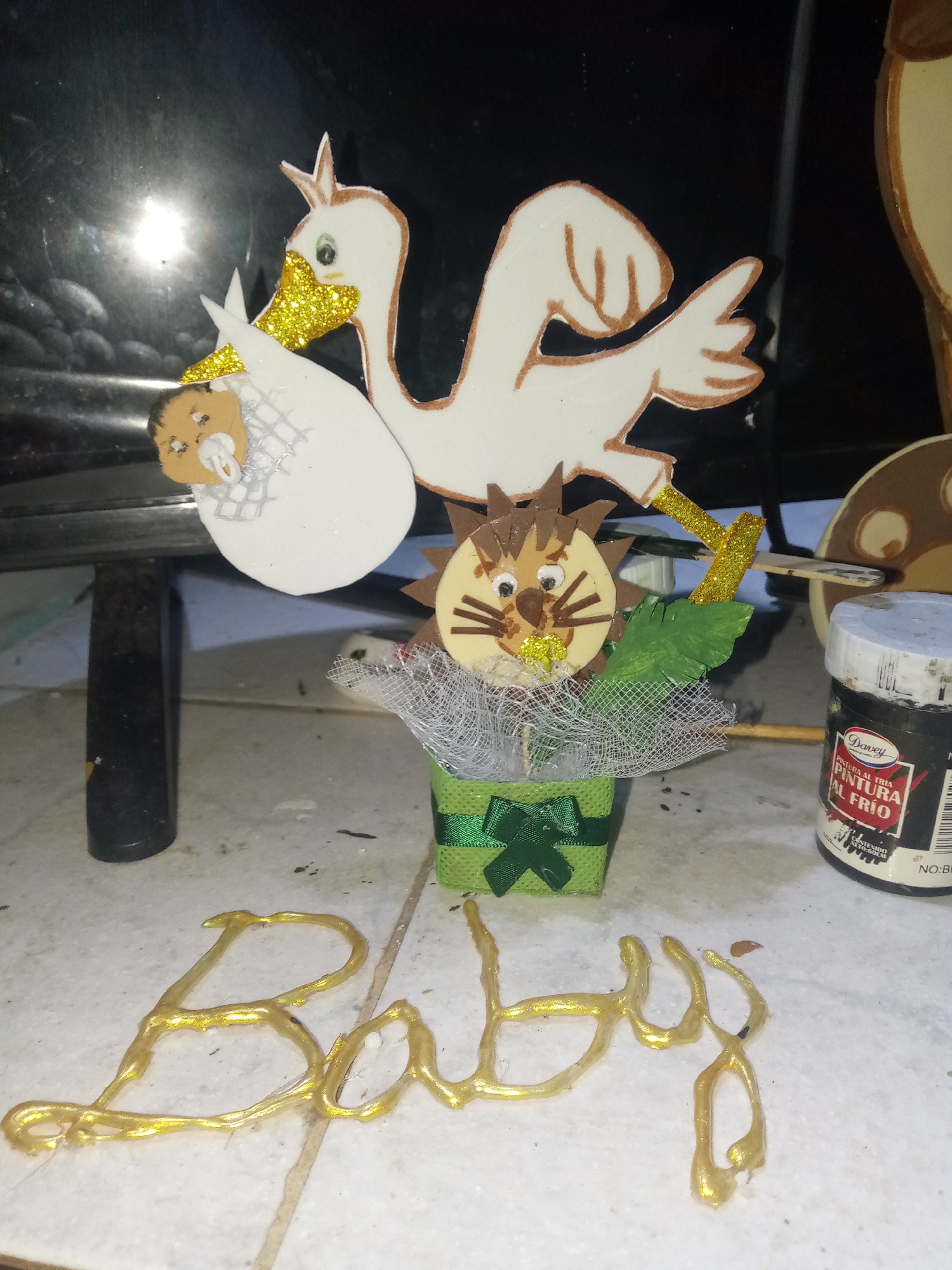 The image features a baby shower decoration set against a blurry background. The decoration includes a stork carrying a baby in a pouch, a lion, and a small green gift box filled with netting and faux leaves. The word "Baby" is spelled out in gold paint on a white tile surface in the foreground. A black paint jar labeled "Davey Pintura al Frío" is visible on the right side of the image. No specific landmarks are visible to confirm the location in Juan Griego, Venezuela, however, the language on the paint jar indicates a potential Spanish-speaking environment.