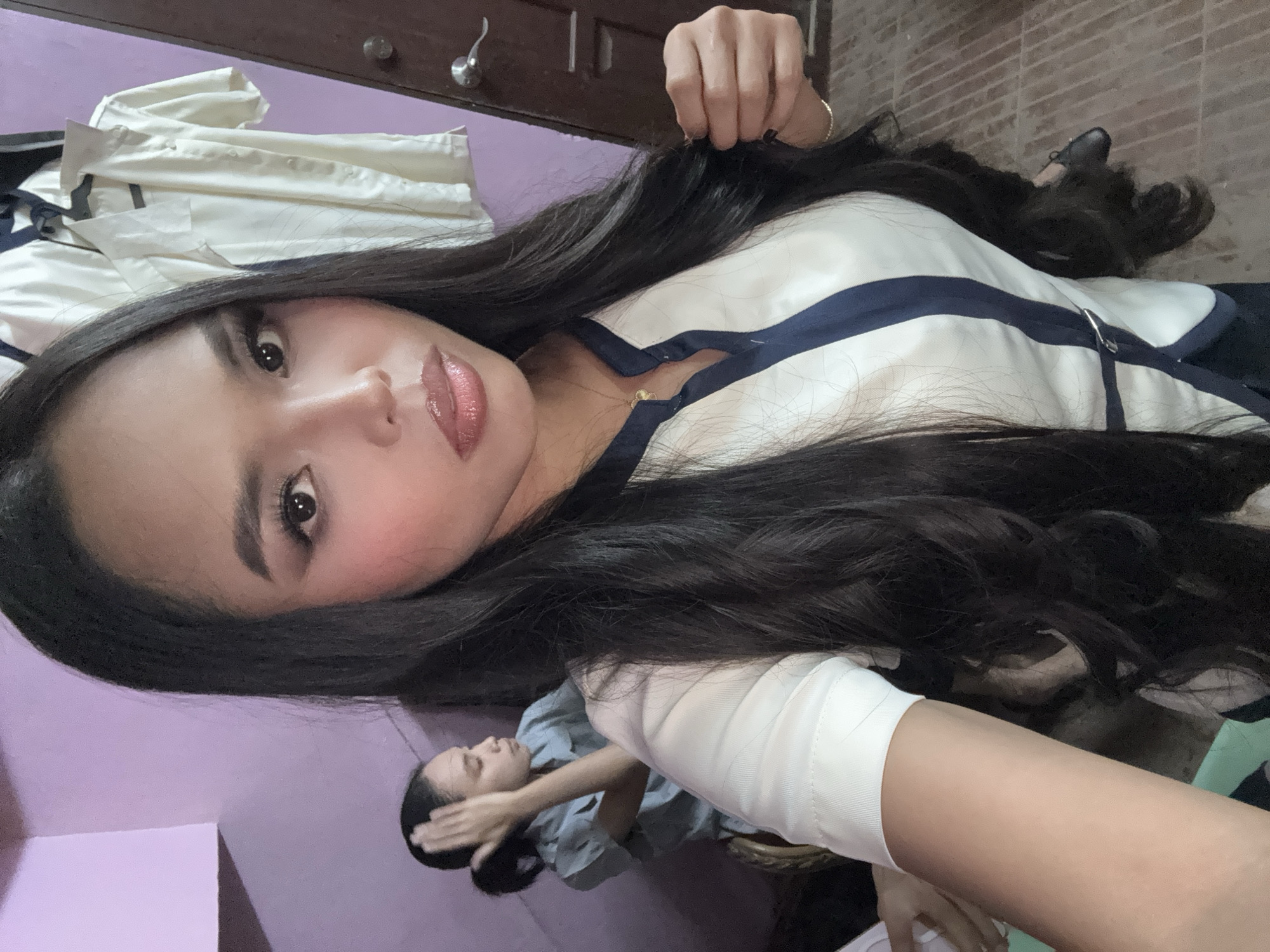 A close-up, selfie-style shot captures a woman with long, dark, wavy hair, wearing a white collared shirt with navy blue trim. She is looking directly at the camera, her right hand is near her hair. She is wearing makeup, including winged eyeliner and lip gloss. A woman with her hair in a ponytail is visible in the background, to the left, touching her head. The background includes a pale purple wall, a brown wooden door with a silver knob, and several white shirts hanging on what appears to be a rack. The floor is tiled in a brick pattern. The image is taken indoors, likely in a residential setting. The presence of uniforms suggests a school or workplace environment. This photo was likely taken in Davao City, Philippines, given the prevalence of similar attire in educational institutions and the general context of the photo.