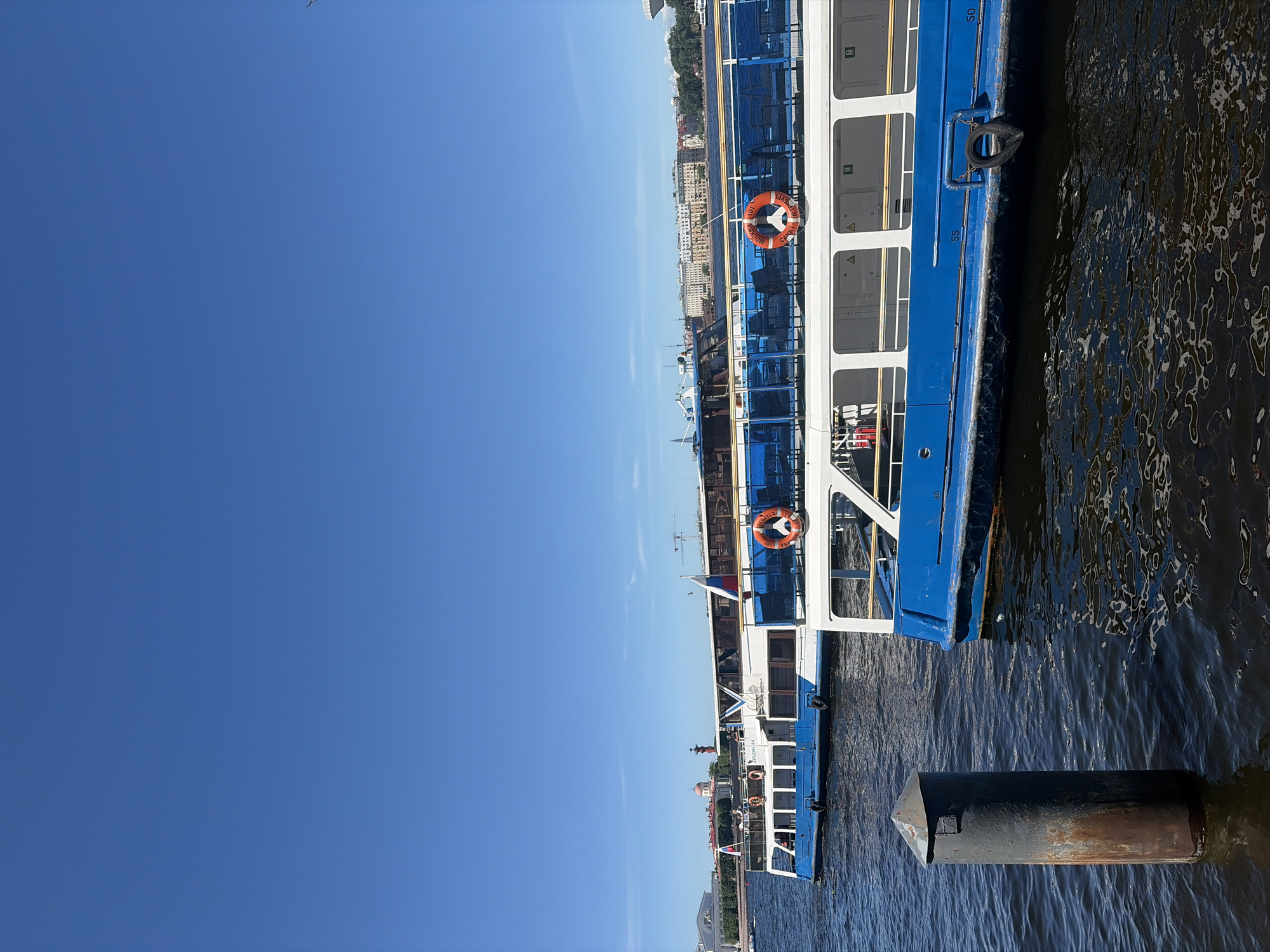 The image captures a sunny daytime scene of passenger boats on a dark blue river, with the cityscape of Saint Petersburg, Russia, visible in the background.
In the foreground, a dark grey, rusty metal bollard with a conical top emerges from the rippled water on the left side. To the right, the side of a blue and white passenger boat is visible, featuring a white superstructure with windows and a blue hull section. A black fender tire hangs from its side near the stern, which bears the number "50."
Beyond this boat, another similar blue and white passenger boat is positioned further back in the water. This vessel is prominently featured, displaying a Russian flag on its stern and a lifebuoy with white text "МОСКВА - 101" and "САНКТ-ПЕТЕРБУРГ" on an orange ring, affirming the location. A few indistinct passengers are visible on its upper deck. Another Russian flag is visible towards its bow.
The background reveals a panoramic view of Saint Petersburg's architectural landmarks along the riverbank. Distinctive pale classical buildings with columns are visible on the left, likely part of the Palace Embankment. Further right, a prominent red-domed building (the Kunstkamera or similar structure) and a red Rostral Column are clearly identifiable on the Spit of Vasilyevsky Island. Beyond these, a continuous line of multi-story buildings with various architectural styles, interspersed with green trees, stretches along the far bank. Construction cranes are also visible in the distance above the buildings.
The sky is a clear, bright blue with only very faint wisps of cirrus clouds, indicating fair weather. The water reflects pockets of sunlight, creating shimmering patterns.