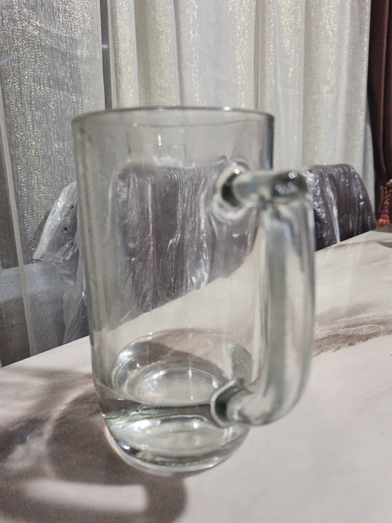 An empty, clear glass mug with a handle sits on a light-colored patterned tablecloth. Behind the mug, sheer curtains with a shimmering pattern are visible. The mug is slightly angled, and there is a small amount of liquid at the bottom. The environment appears to be indoors. The location in the city of Kelapapati, Indonesia is not discernible from the image.
