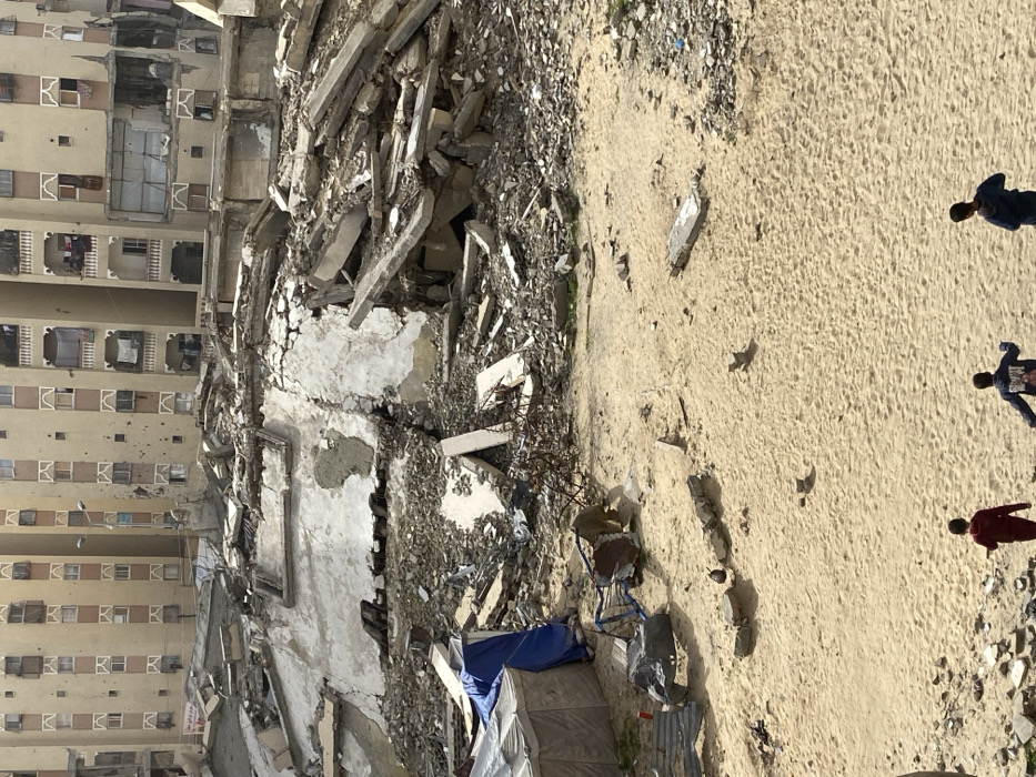 The image displays a scene of urban devastation in Khan Yunis, Palestinian Territory, under daylight conditions. In the foreground, three children walk from left to right across an open, sandy area interspersed with scattered debris, including small concrete fragments. To the left of this sandy expanse, a makeshift camp appears, consisting of a rectangular grey tent and a smaller structure covered by a blue tarp. Nearby, a twisted metal frame, possibly from furniture or a cart, lies on the sand.
The midground is dominated by extensive piles of concrete rubble, broken slabs, and exposed rebar, indicative of a collapsed or severely damaged building. Structural components, including portions of white-painted walls and fractured concrete beams, are clearly visible within the debris field.
In the background, a row of multi-story residential apartment buildings stands. These buildings, appearing light beige or tan, exhibit significant damage, including numerous pockmarks on their facades, broken or missing windowpanes, and sections of balconies either damaged or entirely missing. Some balconies show laundry hanging. A street light pole is visible rising between two of these structures. The sky overhead is clear.