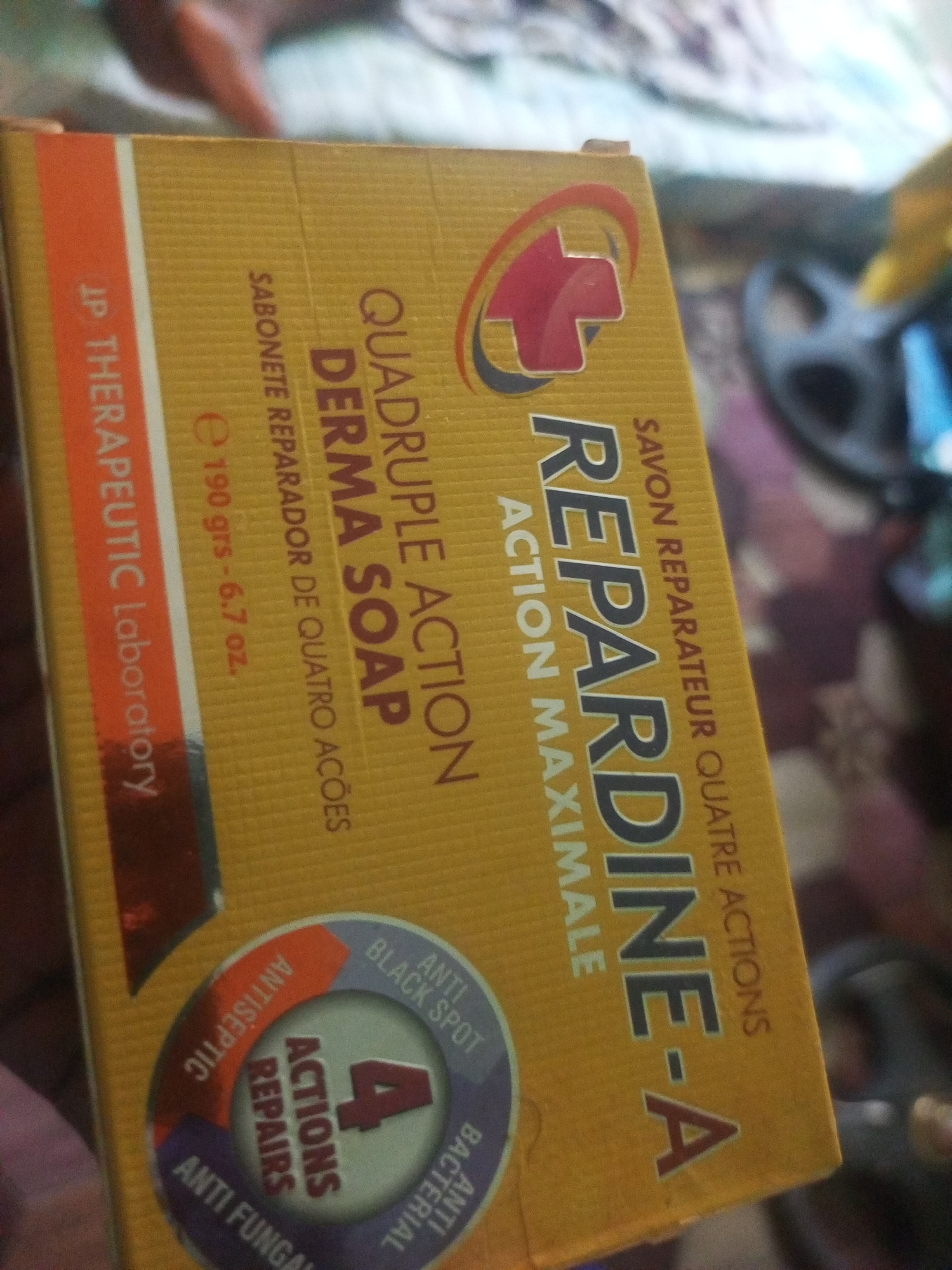 This is a close-up photograph of a yellow bar of soap packaging, likely taken indoors. The packaging is mostly in focus, revealing product information and branding. The soap is labeled "REPARDINE-A" and boasts "QUADRUPLE ACTION DERMA SOAP" with "ACTION MAXIMALE." It also highlights "SAVON REPARATEUR QUATRE ACTIONS" and "SABONETE REPARADOR DE QUATRO AÇÕES," indicating it has four repairing actions. Further details include "190 grs - 6.7 oz." and "JP THERAPEUTIC Laboratory." Circular graphics on the packaging indicate "4 ACTIONS REPAIRS," along with claims of being "ANTISEPTIC," "ANTI BLACK SPOT," "ANTI BACTERIAL," and "ANTI FUNGAL."
The background is blurred and out of focus, showing a person's arm and shoulder in the upper left, suggesting the photograph was taken by someone holding the product. In the background, there are hints of patterned fabric or bedding, and a dark, circular object, possibly a piece of furniture or equipment, is visible on the right. The lighting appears to be indoor ambient light, with no strong indications of the time of day or weather. The overall impression is a casual, everyday scene focused on showcasing the product. The provided location context of Jalingo, Nigeria, is not directly evident from the visual cues within the image itself.
