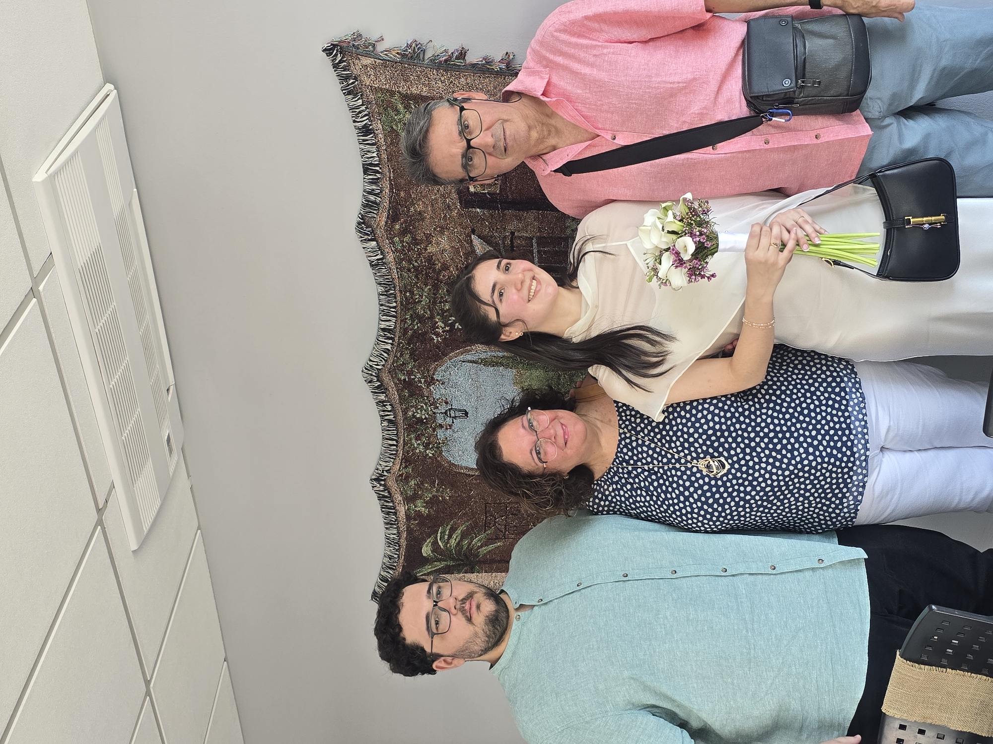 A medium shot captures four individuals posing for a photograph against a wall adorned with a tapestry. The tapestry depicts a pastoral scene with arches, foliage, and a lantern. A white air conditioning unit is visible on the suspended white tiled ceiling.
From left to right, the individuals are:
* A man with dark, curly hair and glasses, wearing a light blue collared shirt. He stands with his arms at his sides.
* A woman with dark, curly hair and glasses, smiling and wearing a navy blue and white polka-dot top.
* A young woman with long, dark hair, smiling and wearing a light-colored, sleeveless dress. She holds a bouquet of white calla lilies and purple flowers, and carries a black handbag. Her left hand rests on the shoulder of the man to her right.
* A man with graying hair and glasses, wearing a pink collared shirt. He has a dark shoulder bag across his chest.
The overall lighting suggests an indoor setting, possibly a room in the city of Terrassa, Spain, given the context provided. The individuals appear to be together for a special occasion, suggested by the bouquet and the formal attire of the young woman.