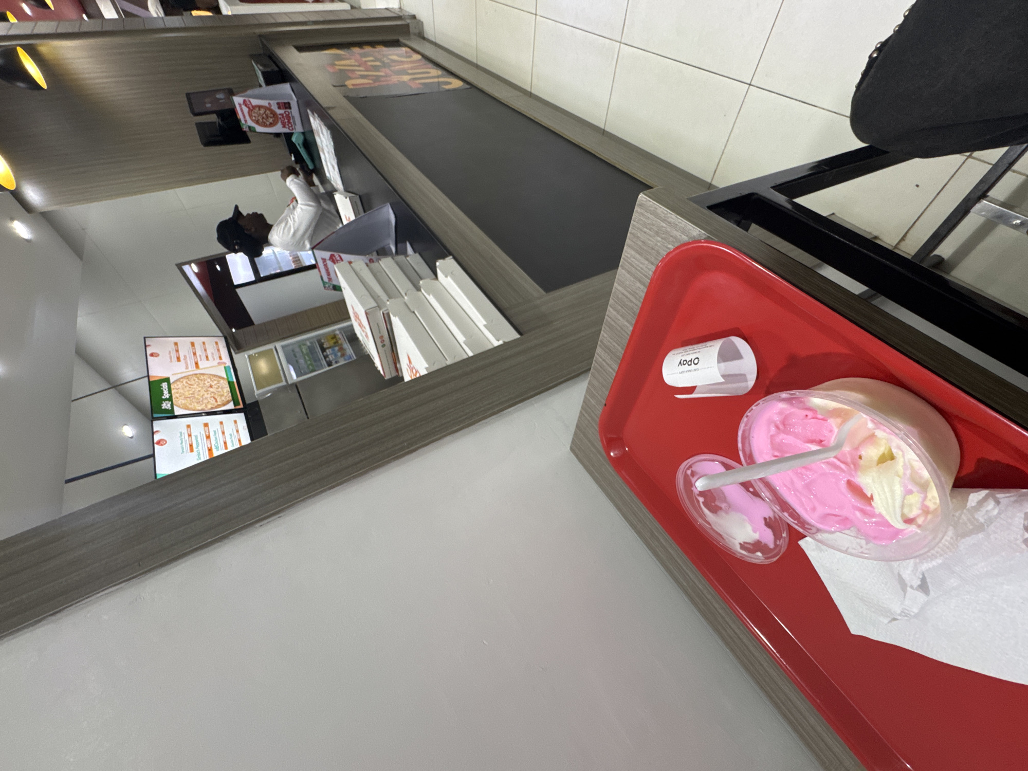 The image, rotated 90 degrees clockwise, captures an indoor scene at what appears to be a fast-food restaurant or a food court.
In the immediate foreground, a red serving tray is placed on a table or counter. It holds a clear plastic cup filled with two flavors of ice cream, pink and a lighter yellow/cream, with a white plastic spoon inserted. A detached lid for the cup rests beside it, along with a crumpled white napkin. A rolled-up paper receipt or strip, clearly displaying the text "OPay", is also on the tray.
Beyond the tray, a long, wooden-patterned counter stretches across the midground. The top surface of this counter is dark and flat. Beneath it, several white cardboard boxes, likely for food items like pizzas, are neatly stacked. One box is partially visible, showing a pizza graphic and the text "MEGA COMBO". Behind the counter, a person wearing a white shirt and a dark cap is visible, seemingly an employee, facing away from the camera. A small monitor or screen is also present on the counter near the employee.
The background features digital menu screens displaying various food options, including images of pizzas. One screen clearly shows the word "Specials". The ceiling with recessed lighting is reflected in what appears to be a mirrored or highly polished surface above the counter area. A portion of a black chair or stool is visible on the far right, suggesting a seating arrangement. The setting implies a casual dining experience, possibly after an order has been placed or received.