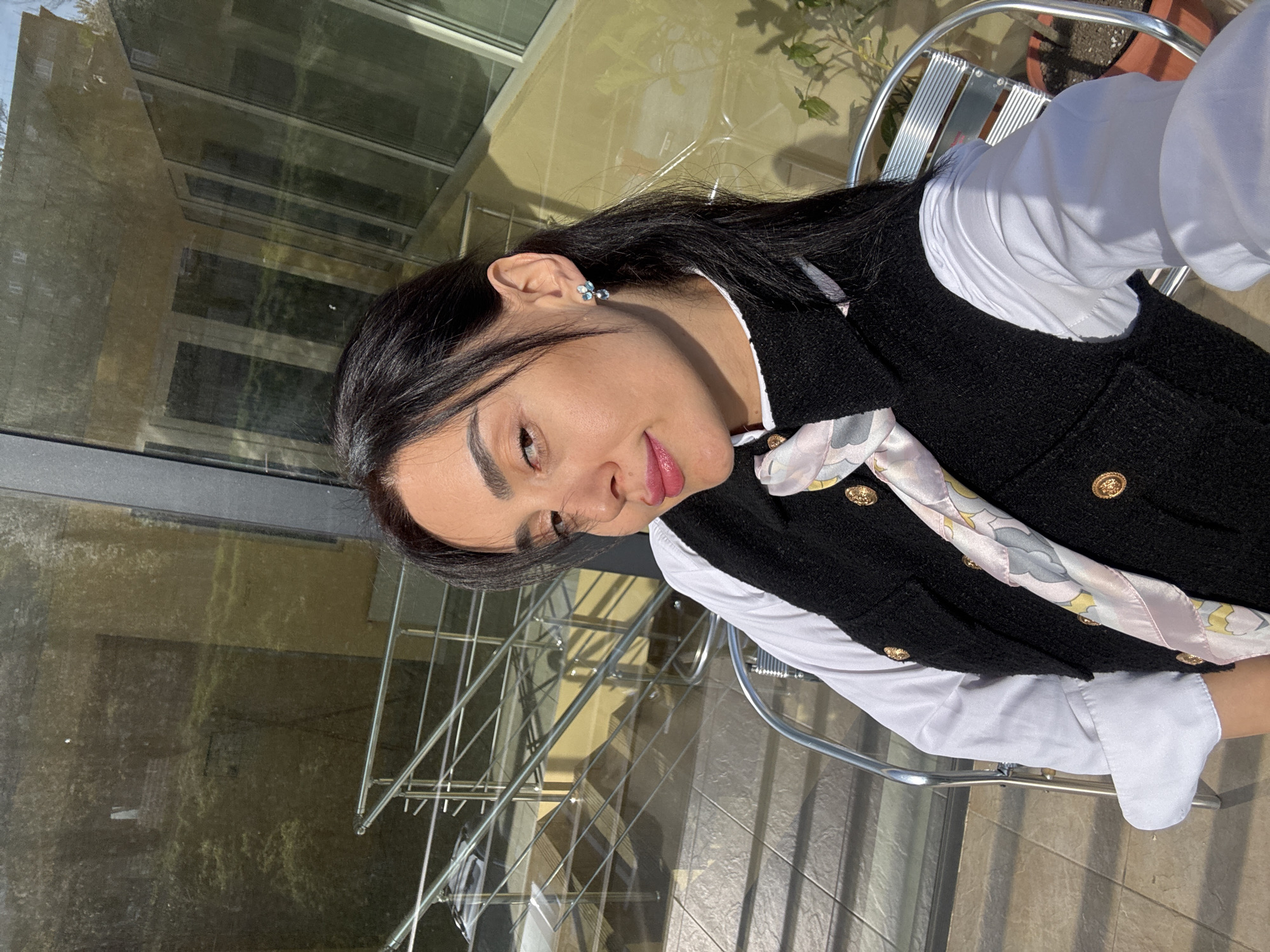 The image, rotated 90 degrees clockwise, presents a woman seated outdoors, bathed in bright natural sunlight, likely on a balcony or terrace in Tbilisi, Georgia. The woman, with dark hair, dark eyes, and a fair complexion, looks directly towards the camera with a calm and gentle expression, her lips slightly parted in a soft smile. She is dressed in a white collared shirt, covered by a black, textured vest featuring several prominent gold buttons. A light-colored scarf, patterned with delicate swirls and floral motifs in shades of pink, yellow, and grey, is tied around her neck. A small, light blue stud earring is visible in her left ear.
The background is dominated by large, reflective glass surfaces, such as windows or panels, which show clear reflections of a multi-story building and hints of greenery, suggesting a modern urban environment. Part of a metal railing and the slatted seat of a metal chair are visible, indicating an outdoor seating area. The ground beneath the chair appears to be tiled. The strong, direct sunlight suggests it is daytime, either morning or afternoon. There is no visible text in the image.