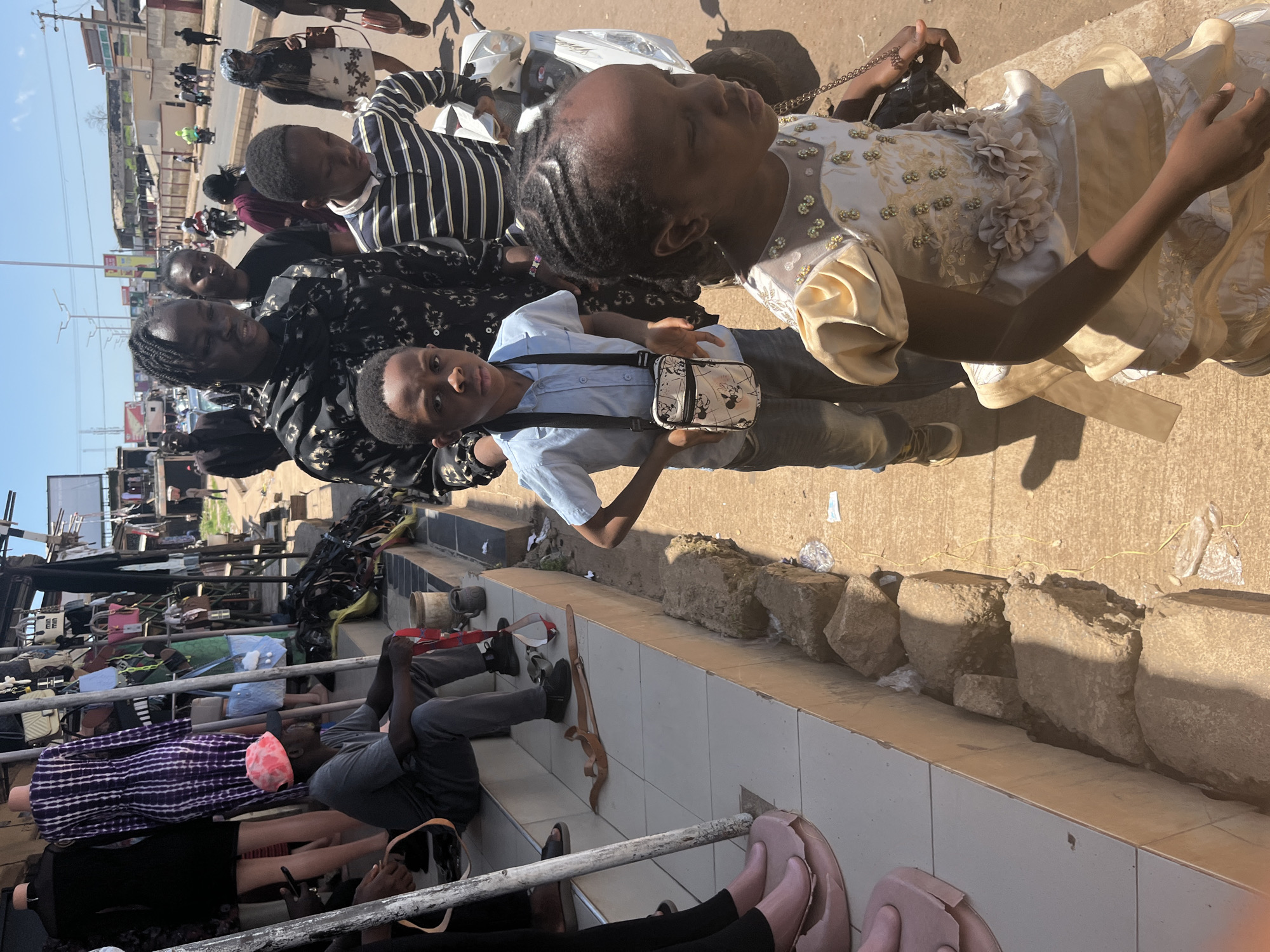 The image captures a street scene in Jos, Nigeria, likely near a market or shopping area. In the foreground, a young boy in a blue shirt and jeans, carrying a small crossbody bag, stands facing forward with a neutral expression. To his right, a young girl in an ornate, gold-colored dress with floral embellishments walks away from the camera. Behind the children, a woman in a black and gold patterned dress is visible, her face turned away.
Further back, near a row of shops displaying clothing and bags on mannequins, a man wearing a gray t-shirt and dark pants sits on steps, looking to his left. A pink and orange hat rests on his head. The environment suggests an outdoor marketplace with a mix of merchandise and people. The ground is paved, and there are piles of rocks and debris along the edge of the walkway. In the background, other individuals and vehicles, including motorcycles, are present, indicating a bustling urban environment. The sky is clear and bright, suggesting daytime.