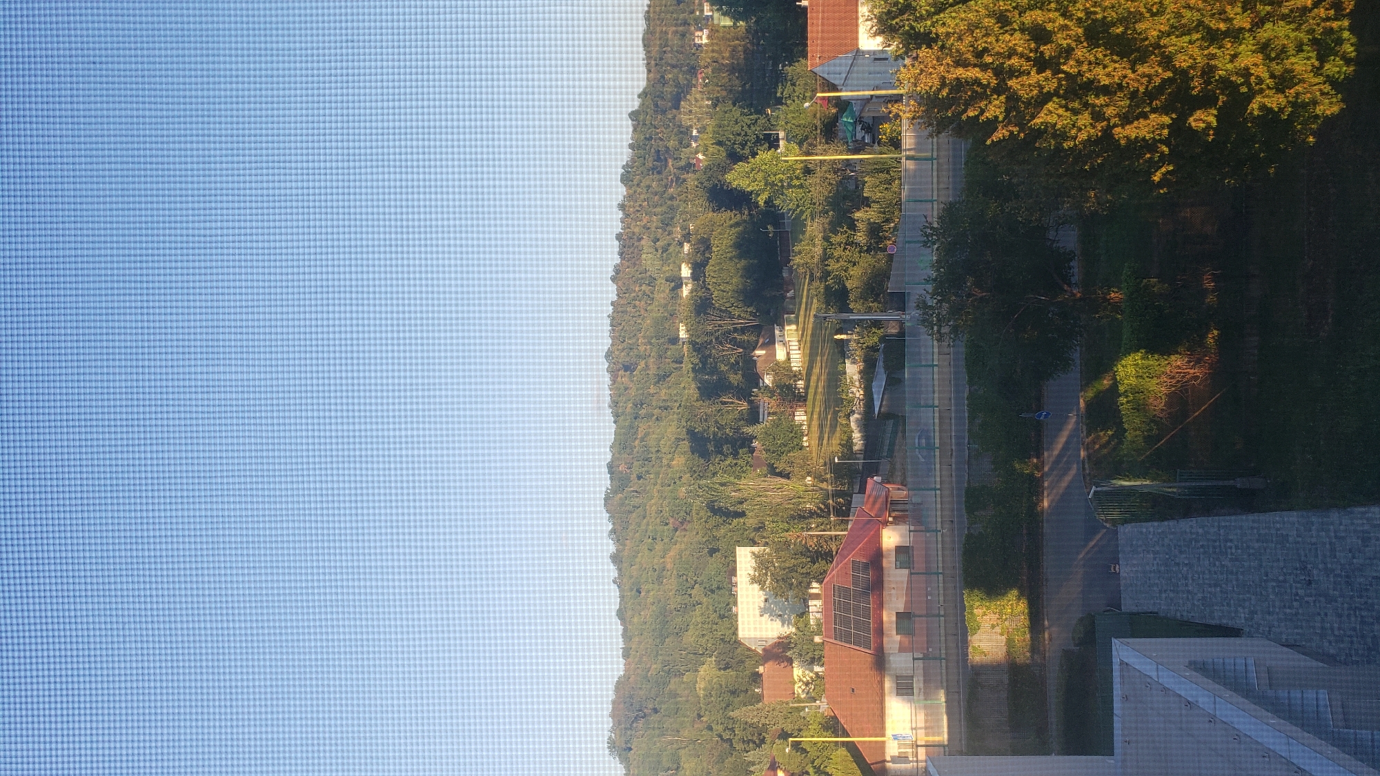 The image shows a view from a window overlooking a residential area in Prešov, Slovakia. In the foreground, a modern building with a gray tiled walkway leads the eye towards a street. Beyond the street, a green fence delineates a grassy area with bushes. Across the street, a row of buildings is visible, including a house with a red tiled roof and solar panels. Further in the background, a dense, tree-covered hill rises, with several scattered houses nestled within the foliage. The sky above is a clear, pale blue, suggesting it is daytime, possibly late afternoon given the warm light illuminating the trees on the right side of the image. The overall impression is of a peaceful suburban landscape.