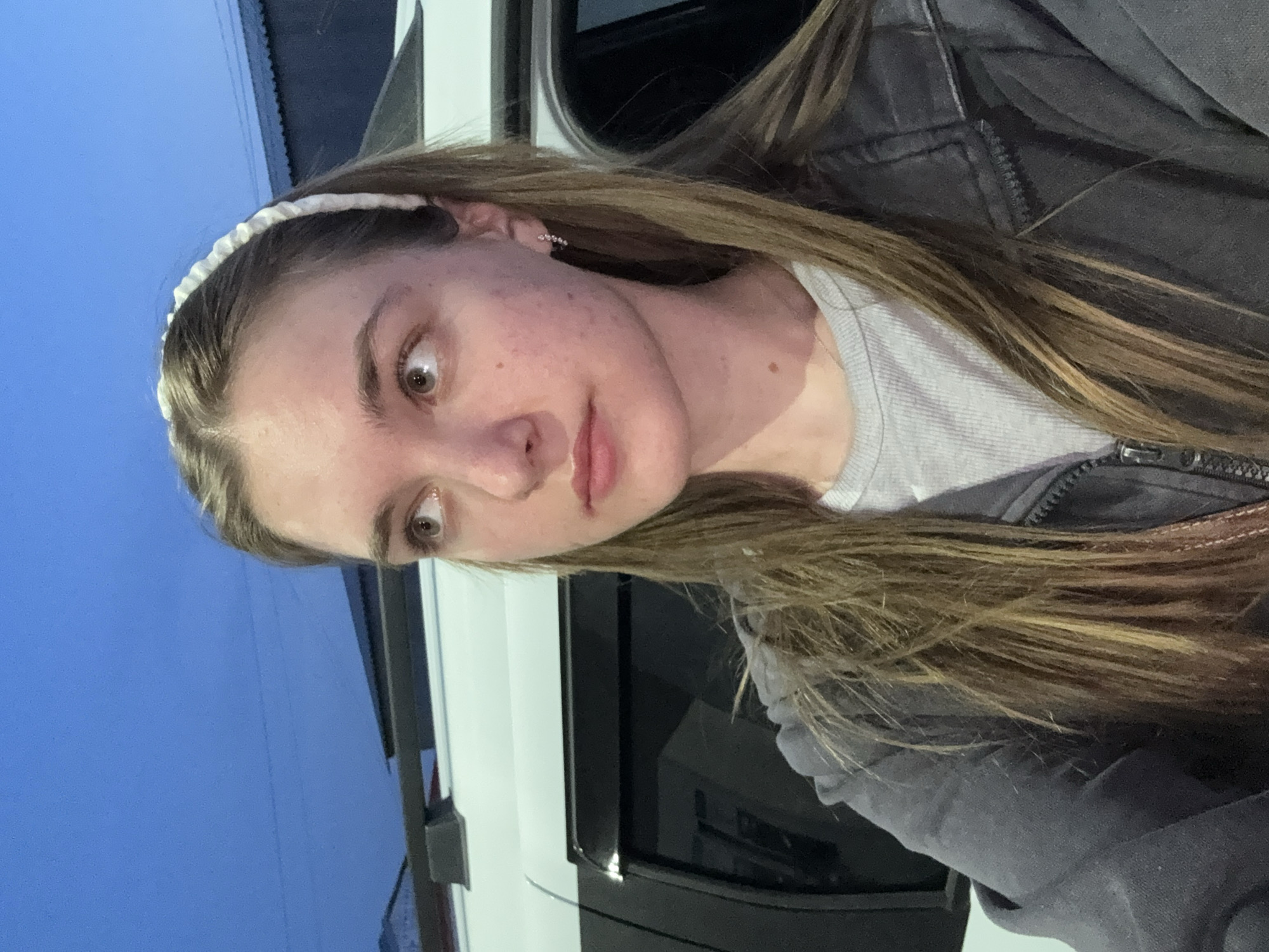 A young woman with light brown hair is standing next to a white vehicle. She is wearing a white headband, a light gray shirt, and a dark gray jacket. The background includes a blue sky and a building. The location cannot be determined from the image.