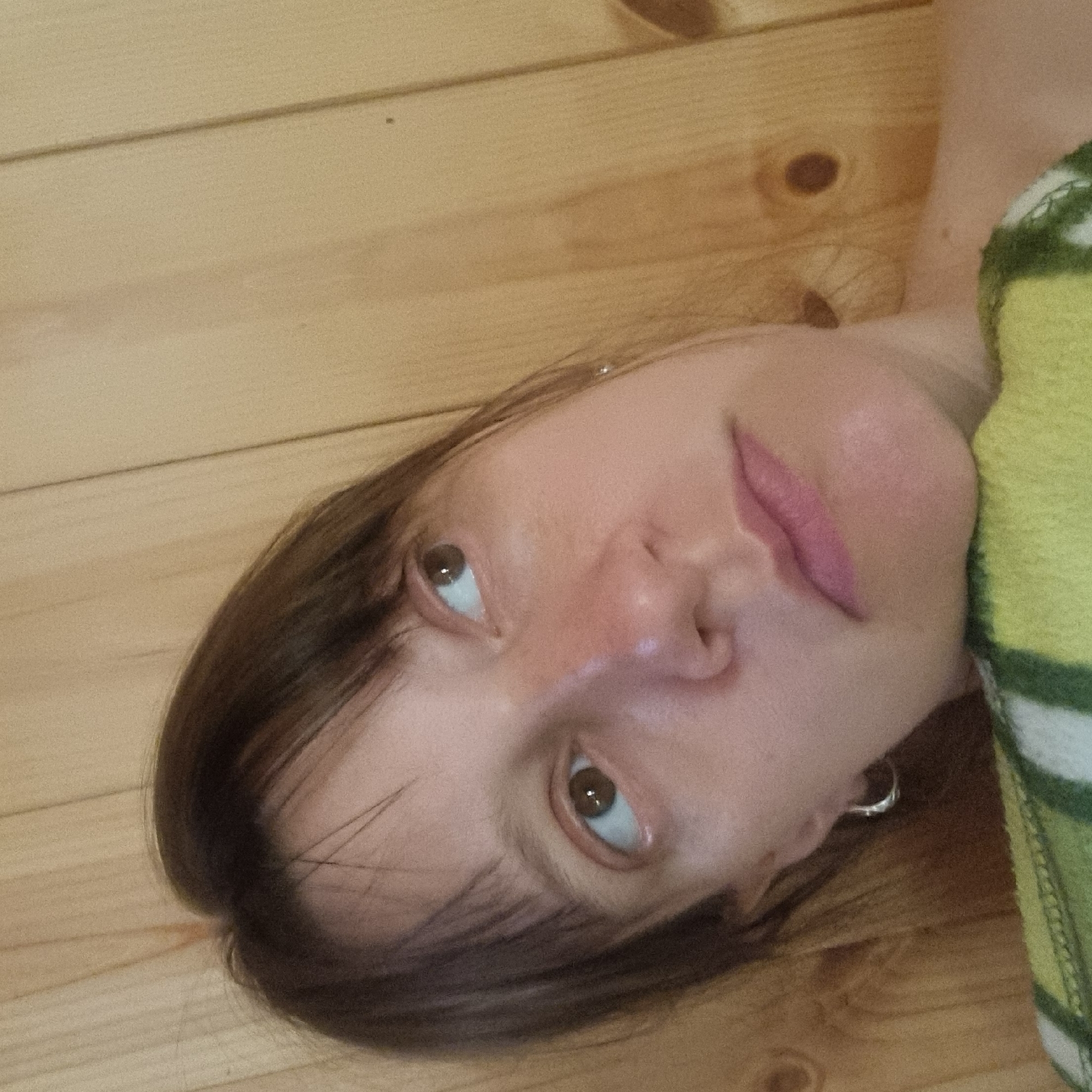 The image shows a close-up, indoor portrait of a person with medium-length brown hair and brown eyes. The person is looking upwards and to the right. They are wearing a silver hoop earring in their left ear. The person's face is partially obscured by a green and white striped fabric, which appears to be a towel or blanket. The background consists of light brown wooden planks, suggesting an indoor environment with wooden paneling. There are no discernible landmarks or contextual clues that would allow for the identification of a specific location within Dunnae-myeon, South Korea.