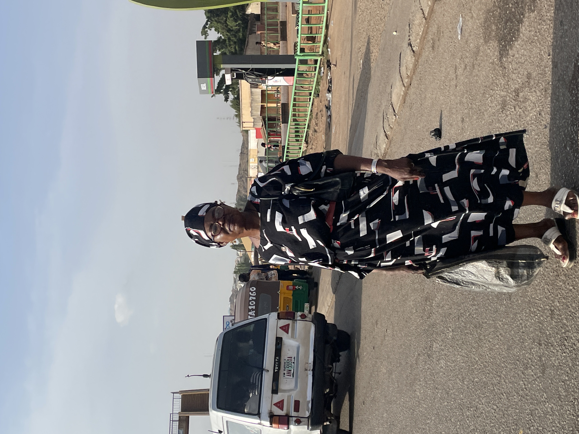 The image shows a woman standing on a street in Jos, Nigeria. She is wearing a black and white patterned dress and a headscarf. She is holding a dark-colored bag. Behind her is a white Toyota Hiace van with the license plate "ANW 555 XA." Other vehicles, including tricycles, are visible in the background. A petrol station is also visible in the background, with a green-painted ladder. The sky is light blue with a few clouds. The location appears to be a busy street in Jos.