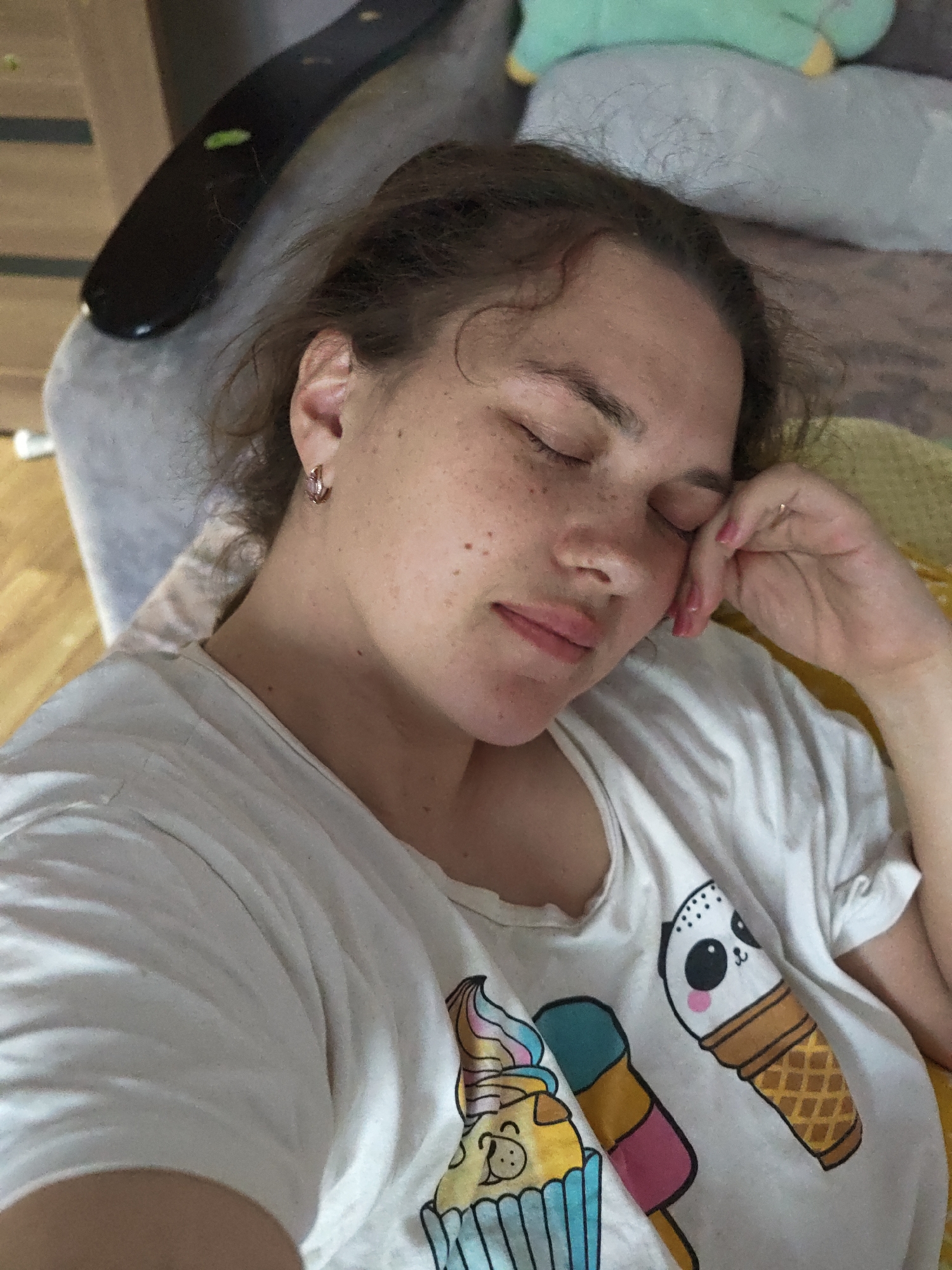 A woman is sleeping. She lies with her eyes closed, her head resting on her hand. She has freckles on her face. She is wearing a white t-shirt with ice cream cone designs. The background includes a light gray surface and a wooden floor. The image was taken in a location with a home-like atmosphere. There is no information available to determine if this image was taken in the city of Chaikovsky, Russia.