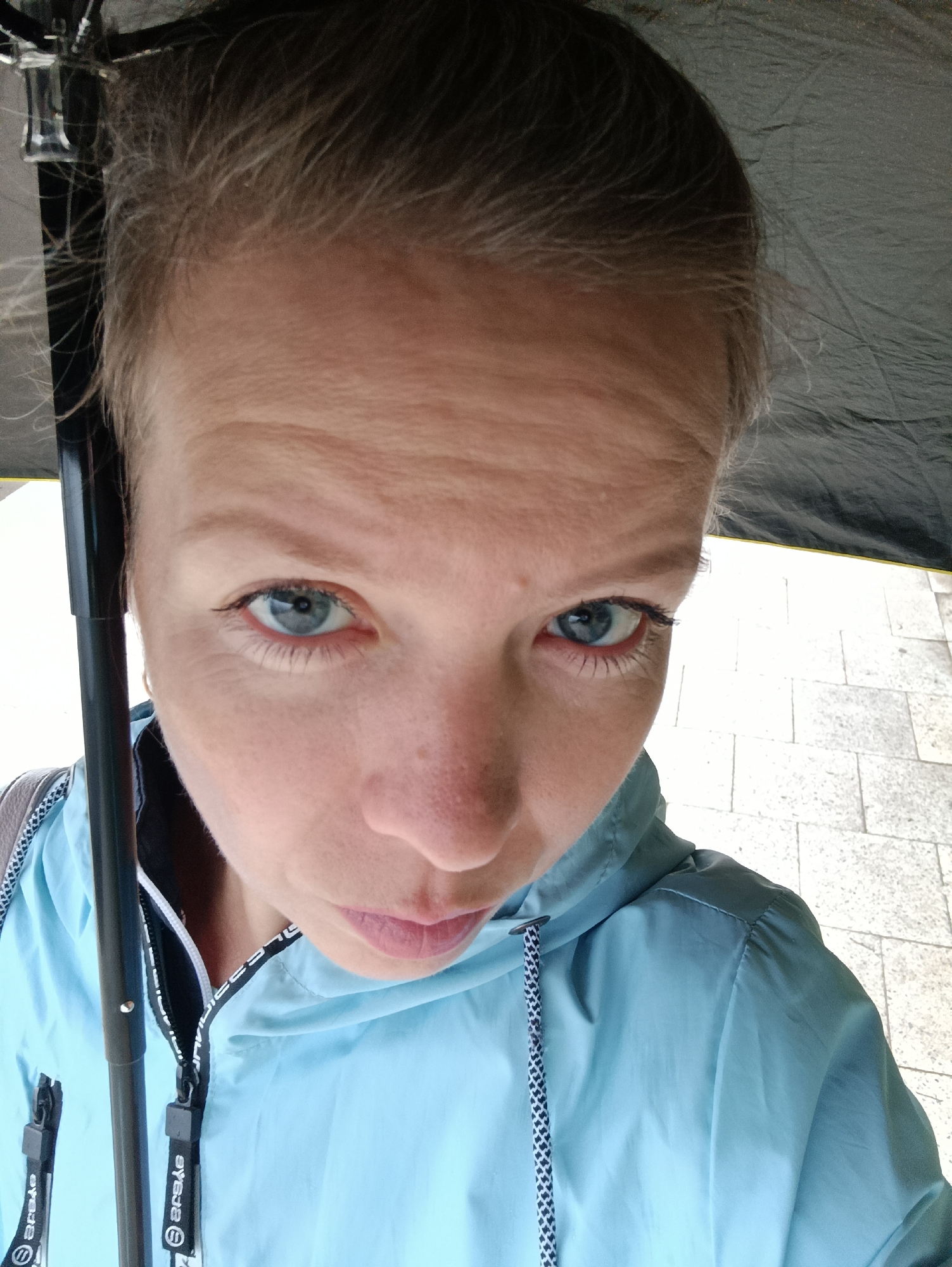 A fair-skinned woman is holding an umbrella while looking at the camera in the city of Chaikovsky, Russia. She has light-colored hair pulled back from her face and light blue eyes. She is wearing a light blue hooded jacket with black and white detailing. The umbrella is black, and some metal parts of the umbrella can be seen in the upper left corner. The background shows light-colored pavement stones.