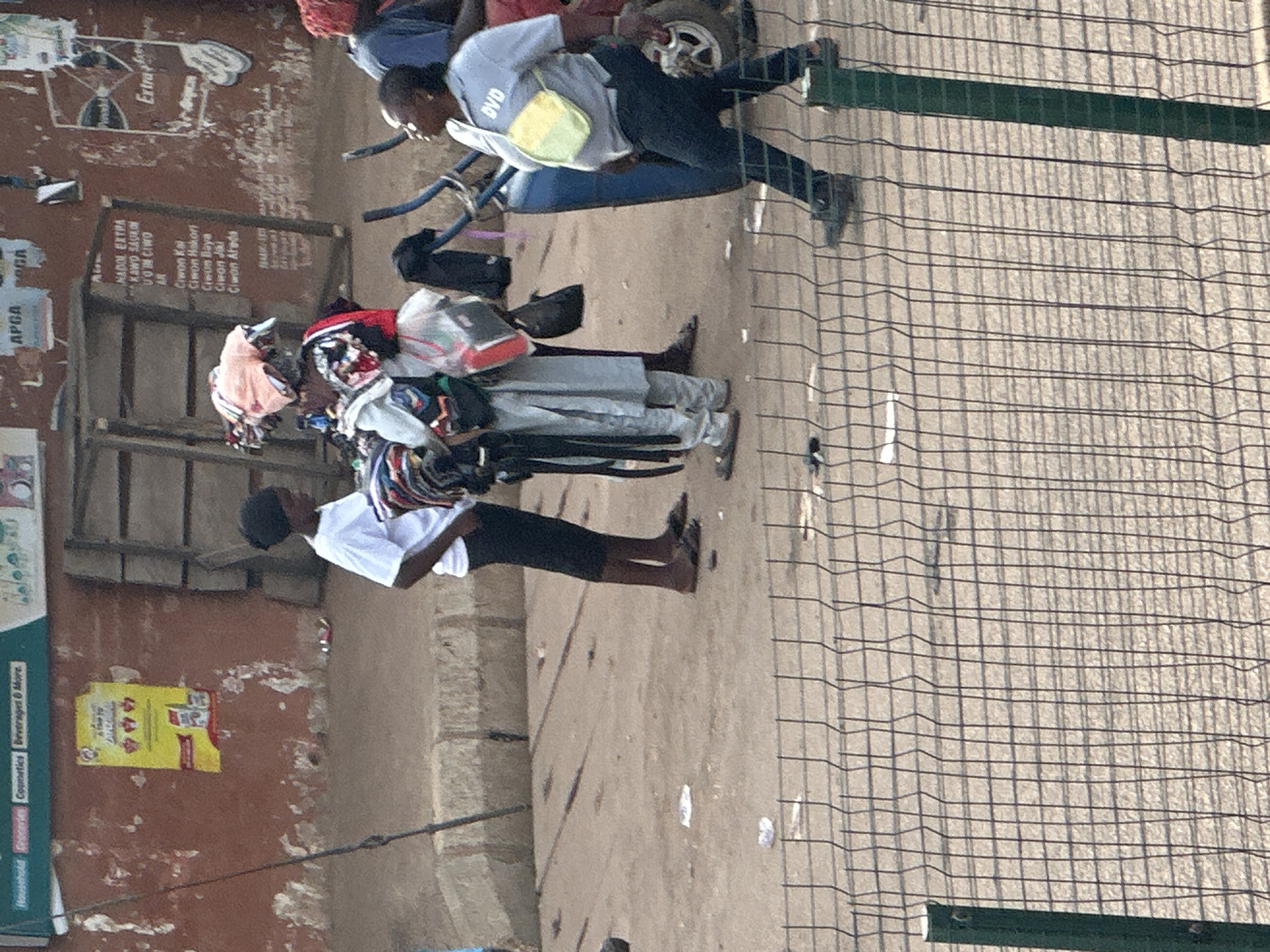 The image depicts an outdoor scene in Bukuru, Nigeria, likely during the daytime under clear skies, as suggested by the bright lighting. The central focus is on several individuals, seemingly engaged in a commercial activity, possibly a street market.
In the foreground, a person wearing a white t-shirt and black shorts stands facing away from the camera, their back to a rough, textured wall. Next to them, a second person, also seen from behind and partially from the side, is dressed in gray clothing and appears to be interacting with a stack of bundled items. These bundles are comprised of textiles, with visible colors and patterns suggesting clothing or fabric.
Further back, another individual in a gray t-shirt with "DVD" printed on it, and dark trousers, is partially visible, appearing to be either a vendor or a customer. They are seated on a blue structure, and a bicycle wheel can be seen behind them. The general setting appears to be a busy street or market area, characterized by the presence of wooden structures, a rough-surfaced ground, and a wire fence in the right portion of the image.
Several signs with text are visible. One sign on the wall contains a list of names or items in what appears to be a local language, including "NADOL EXTPA," "VON CIWO," "AR," "Ciwon Kal," "Cimon Hakorn," "Creon Baya," "Ciwon Jiki," and "Clwon Aheda." Another sign on a vertical surface to the left is partially obscured but includes text like "Cosmetics Beverages & More." A yellow sign with red text and some icons is also visible on the wall, adding to the market atmosphere. The overall impression is of a vibrant, everyday scene in a Nigerian commercial hub.
