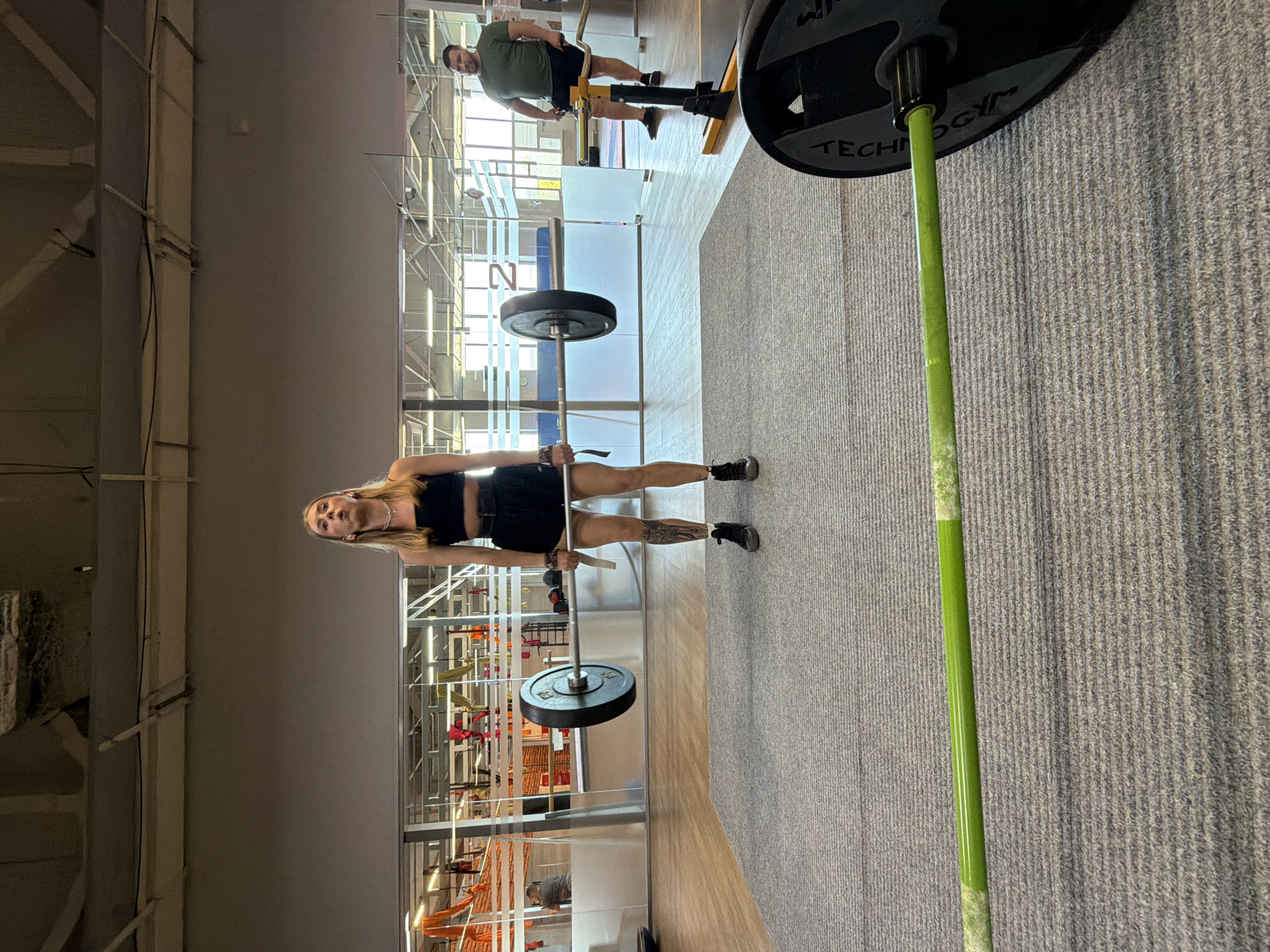 A woman in a black sports bra and shorts is lifting a barbell in a gym. She stands with feet shoulder-width apart, knees slightly bent, and arms extended downward, gripping the barbell in front of her. A tattoo is visible on her left leg. The gym has large windows with a view of what appears to be interior elements of a building with orange and red accents. In the background, a man in a green shirt stands near weightlifting equipment. A textured gray carpet covers part of the floor, and a wooden floor is also visible. In the foreground, part of a green barbell is visible. The location of the city Chaikovsky, Russia cannot be verified from the given image.