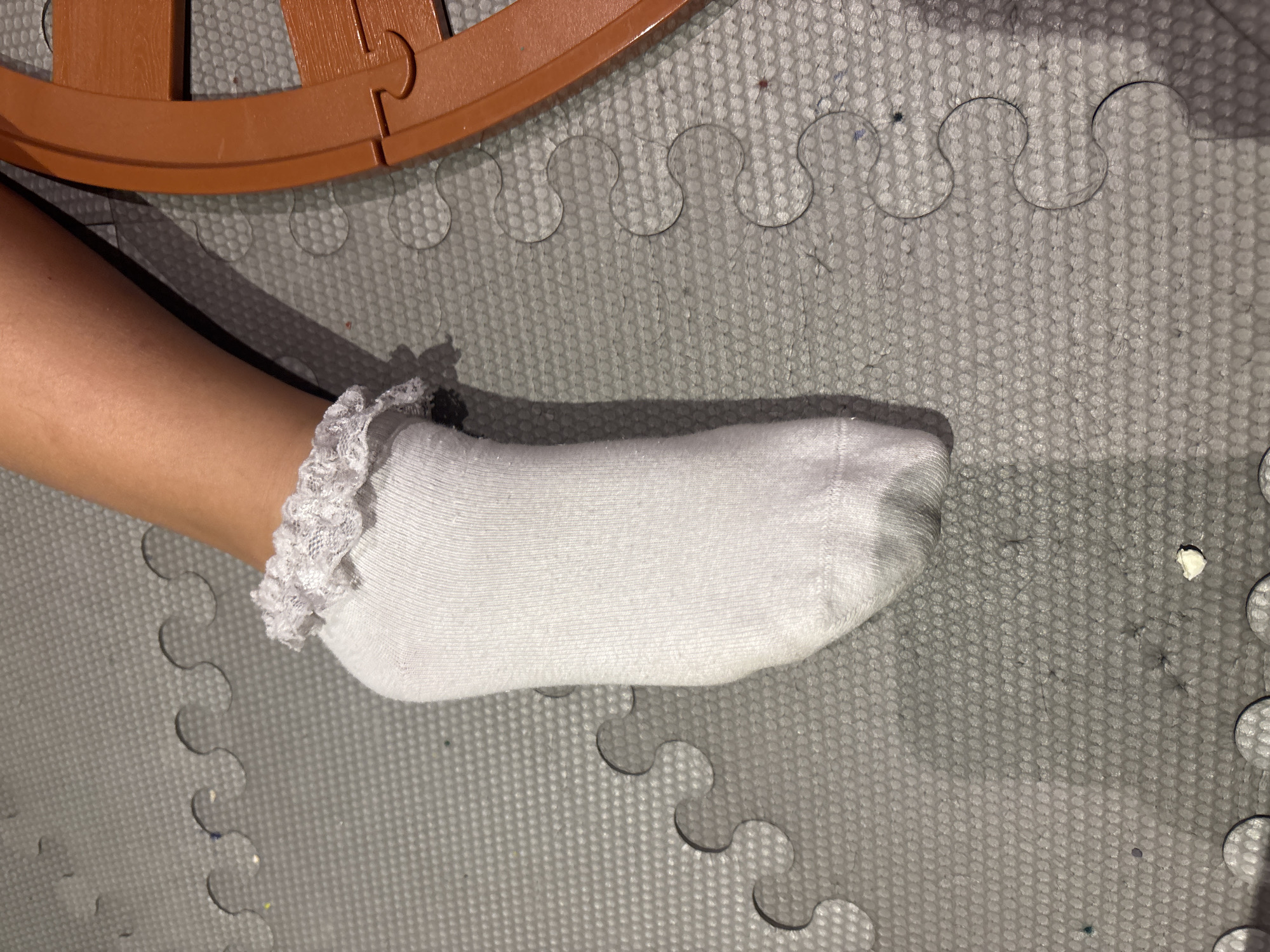 A close-up, high-angle view captures a partial human leg and foot, likely belonging to a child, wearing a white ankle sock with a ruffled lace cuff. The skin tone of the leg is light brown. The foot, encased in the sock, rests on a grey interlocking foam playmat, which features a repeating raised dot texture. Visible interlocking seams of the foam mat appear as wavy lines across the surface. In the upper right quadrant, a curved segment of an orange plastic object, resembling a toy train track piece or a similar interlocking play component, is present. Scattered across the foam mat are small specks of debris and lint. A small, irregularly shaped white fragment is visible on the mat in the lower right area. The specific location in Dubai, United Arab Emirates, cannot be determined from the visual information provided in the image.