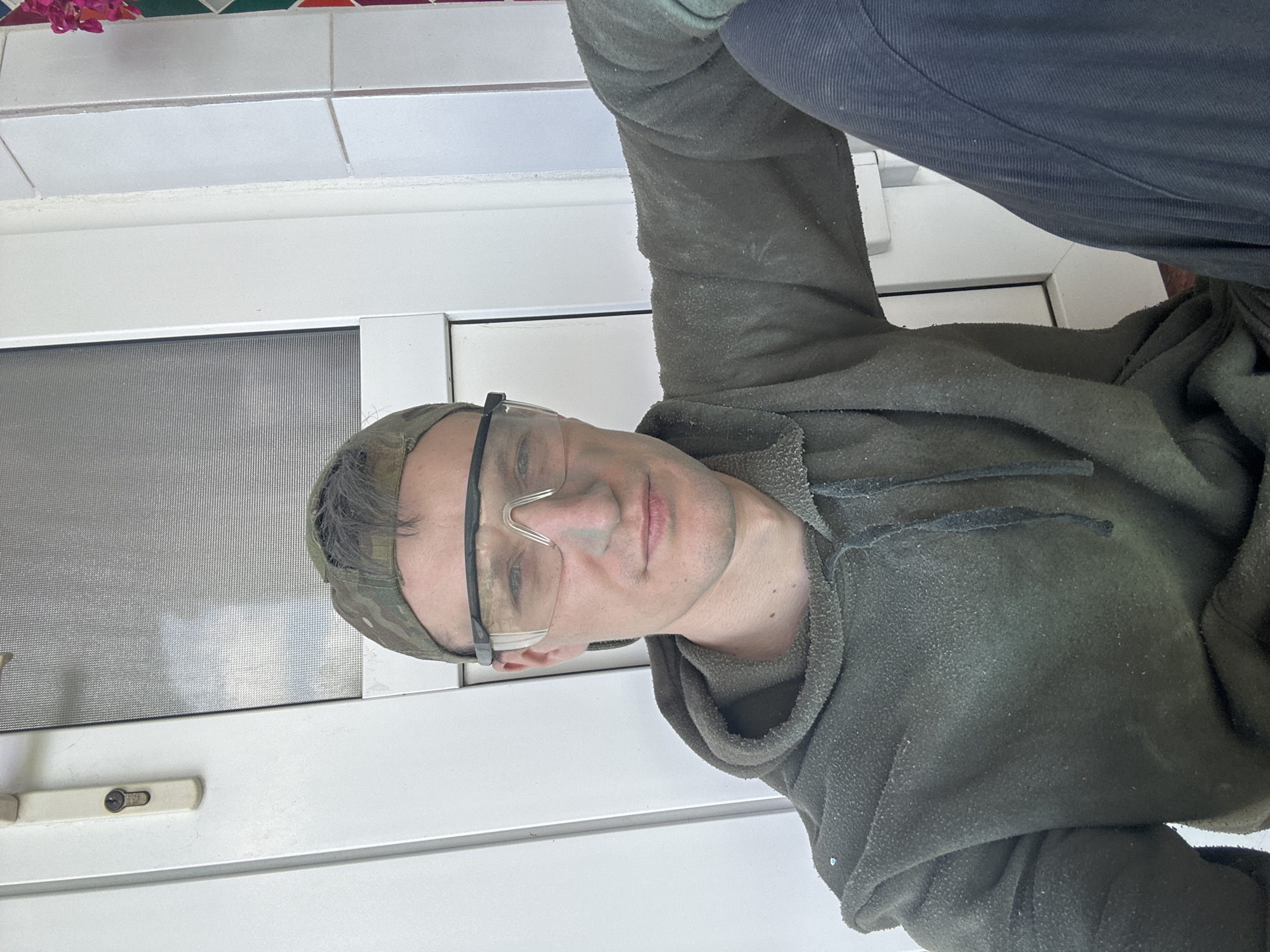 A man wearing a camouflage baseball cap and clear safety glasses is taking a selfie. He is dressed in a dark green hooded sweatshirt and dark pants. He is leaning against a white door frame that has a silver handle and a lock. The background appears to be an outdoor setting, possibly in the city of Makiv, Ukraine, with a tiled wall and a glimpse of pink flowers to the right. The man's clothing is speckled with what appears to be dust or debris.