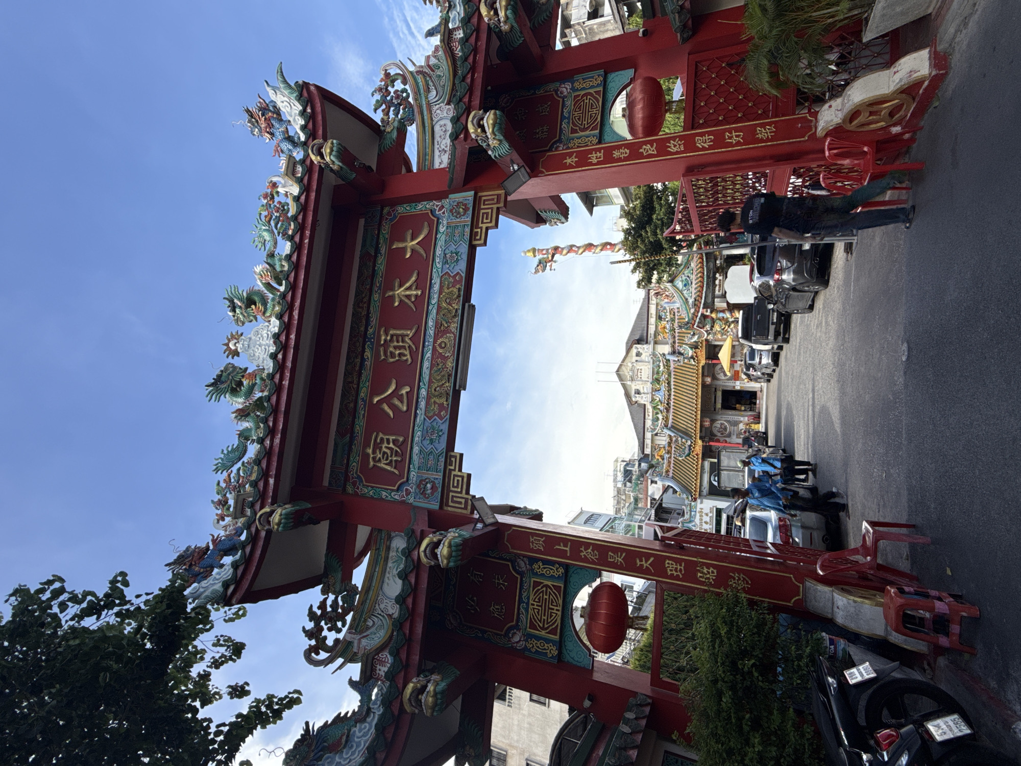 The image depicts a vibrant, ornately decorated Chinese temple gate in Bangkok, Thailand. The gate, predominantly red with intricate green and gold details, is adorned with traditional Chinese motifs like dragons and auspicious symbols. Calligraphy in Chinese characters is prominently displayed on the archway.
Beyond the gate, a street scene unfolds, with cars parked along the sides and people moving about. In the background, a temple structure is visible, with its roof and decorative elements contributing to the overall cultural atmosphere. The sky above is a clear blue.