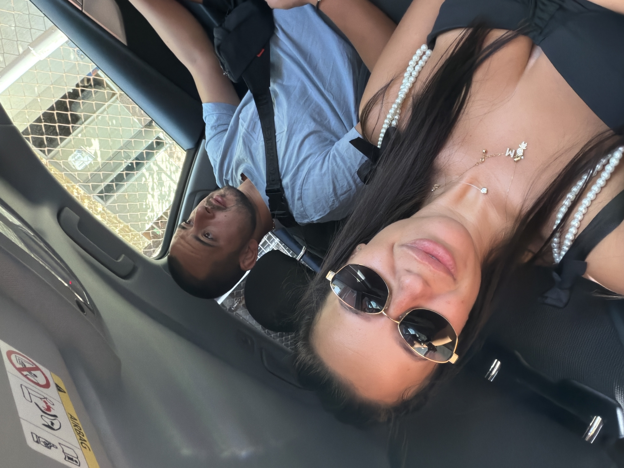 In a car interior, a couple is visible. The woman in the foreground, wearing sunglasses and a black top adorned with pearls, is taking a selfie. She has long, dark hair. Behind her, a man with short dark hair, wearing a light blue t-shirt, is in the driver's seat. He has a dark bag slung across his chest. The car's interior, including an airbag warning sticker on the ceiling, is also depicted. Through the car's windows, a building with a chain-link fence is visible, suggesting an urban setting. The scene appears to be captured during daylight. The location is identified as Burgas, Bulgaria.
