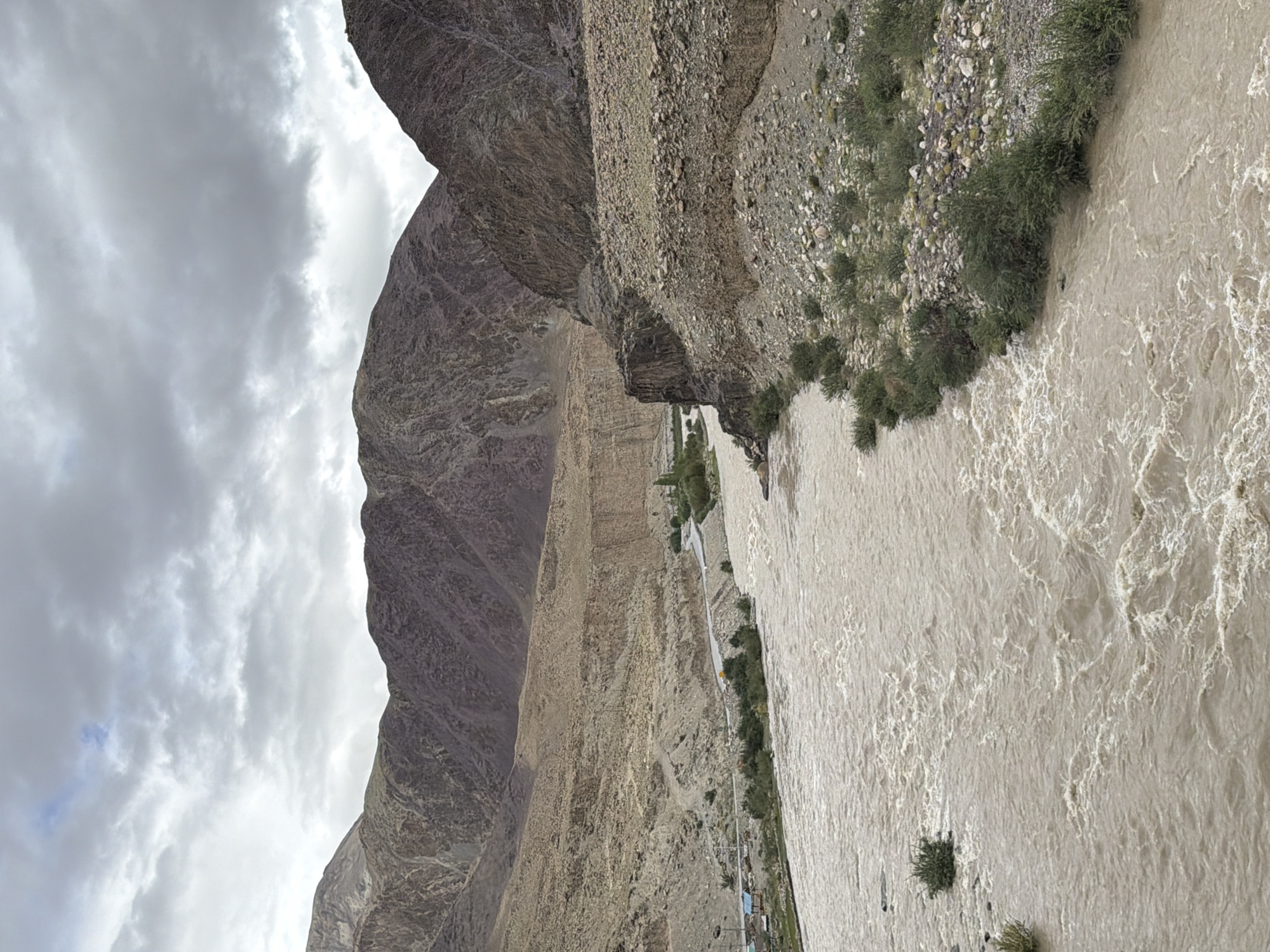 This is a wide-angle, eye-level shot of a muddy, fast-flowing river cutting through a rugged, mountainous landscape. The mountains on either side of the river are steep, barren, and rocky, with slopes showing signs of erosion. The river dominates the foreground and mid-ground, its surface churned with whitecaps and eddies, indicating a strong current. The water color suggests it is carrying a significant amount of sediment, likely from snowmelt or recent rainfall in the higher elevations.
On the left bank of the river, in the mid-ground, there is a small, paved road that winds its way along the base of the mountain. A few scattered buildings, possibly a small settlement or roadside facilities, are visible along this road, along with some trees and green vegetation. On the right bank, the terrain is steeper and rockier, with sparse scrubby vegetation clinging to the slopes.
The sky above is overcast with thick, dark clouds, casting a diffused light over the scene and contributing to a somewhat dramatic and moody atmosphere. A single, small patch of lighter sky with a hint of blue is visible in the upper left quadrant, suggesting a break in the clouds. The overall impression is one of a remote, wild, and powerful natural environment. The image appears to depict the Indus River near Kiari, India, a region known for its dramatic mountain scenery and glacial rivers. There are no people visible in the image.